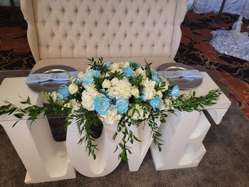 Main table center piece blue and white flowers - A lush, low centerpiece of white hydrangeas and roses punctuated with vibrant blue roses creates an elegant focal point for a wedding or reception table. This flower arrangement for a main table or head table brings a soft blue-and-white palette that reads formal and celebratory-perfect for a wedding, anniversary banquet, or milestone dinner where the person you're honoring should feel truly seen.  Dense clusters of white hydrangea and cream roses form the base while bright blue roses and trailing greenery spill gently across the tabletop, spreading broadly to fill the space between charger plates and folded napkins. The composition sits low for easy conversation, pairing beautifully with decorative linens, charger plates, and large table accents to create a cohesive, photographed-ready display.  Designed to dress a decorated table, this centerpiece works equally well for long banquet tables or a round sweetheart table; it sends a clear message of refined celebration to your guest of honor. Because it's arranged low and full, it's easy to place amid place settings without blocking sightlines-order in advance to ensure availability for your event date.