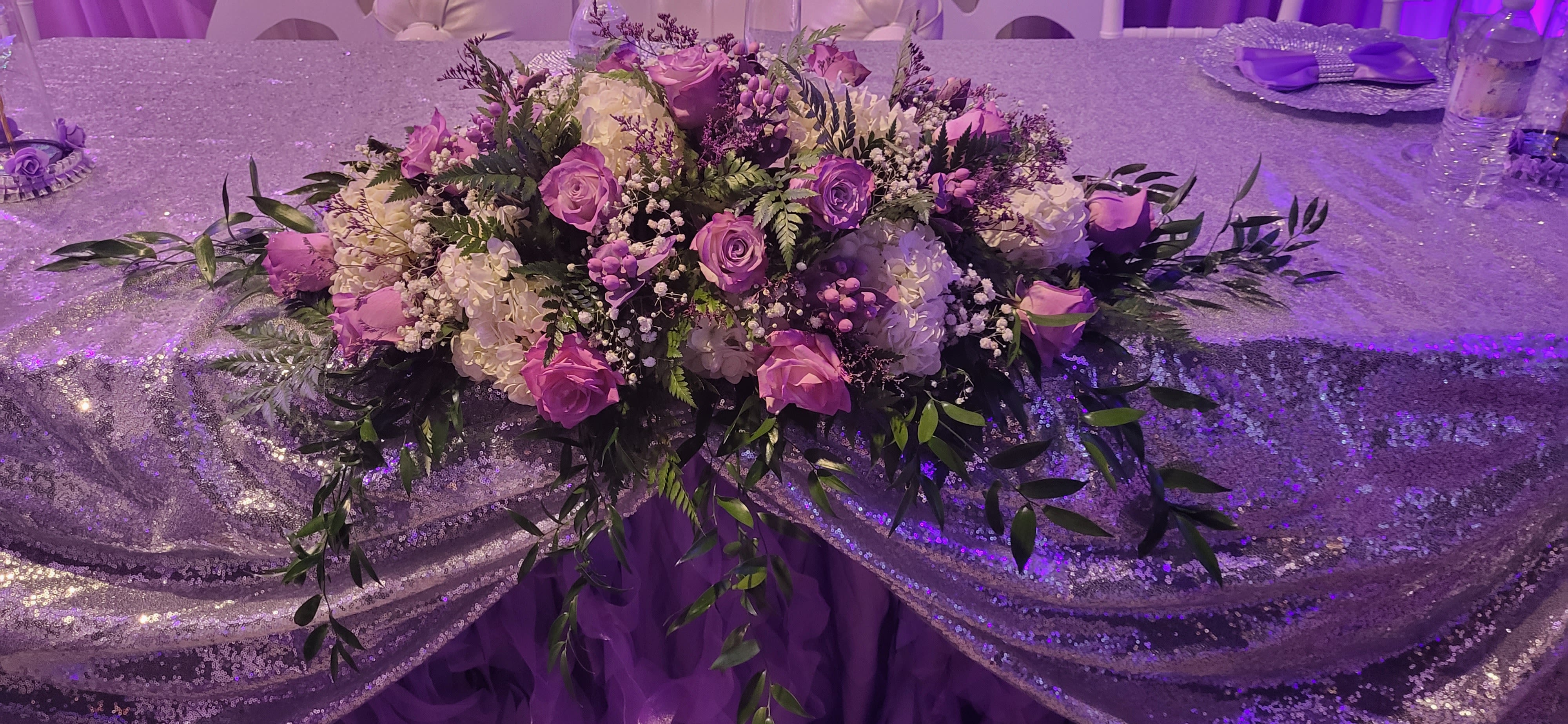 Main center piece for special events purple rose, white hydrangeas  - Lavender roses and cloudlike white hydrangeas form a low, wide centerpiece designed to make guests feel gently celebrated at a wedding or special event. This special events flower arrangement reads as a formal table centerpiece for anniversaries, rehearsal dinners, or elegant receptions-she'll see the romantic purple and white palette and know the moment was planned just for her.  The arrangement features lavender roses, white hydrangeas, and delicate clusters of baby's breath set in a lush, cascading shape with trailing greenery that spills outward from the base. Soft pink and green accents lift the palette, and the low profile keeps sightlines open while filling the table with texture and a subtle floral scent. Purple evening lighting in the venue will deepen the lavender tones for dramatic effect.  Designed to sit on a tablecloth as the focal point, this centerpiece is sized to anchor guest tables without blocking conversation. It's the kind of arrangement that makes someone feel truly seen on their big night-order with confidence for weddings or elegant celebrations to create that intimate, curated atmosphere.