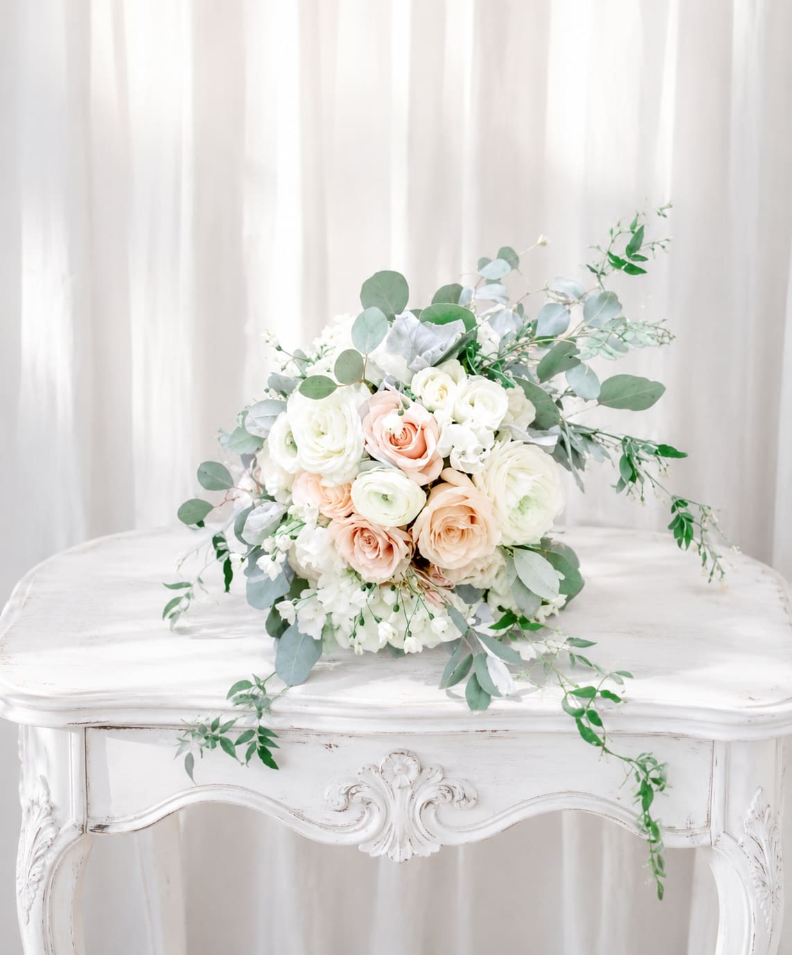 Just for her Bouquet - A pale, romantic wedding bouquet of blush roses and ivory ranunculus that reads like  thoughtful arrangement for a bride or special-occasion recipient. The soft white, pink, and green palette and airy eucalyptus trails give the person you're thinking of an elegant, light feeling; she'll feel genuinely celebrated and seen when this arrives.  This arrangement pairs rounded, soft-headed roses and layered ranunculus with delicate white filler blossoms for texture, all set against loose, trailing eucalyptus that adds movement and a fresh herbal scent. The overall shape is gently rounded with cascading greenery, styled on a white table for a clean, timeless presentation. The combination of blush and ivory tones creates a versatile look that flatters both intimate moments and formal events.  Designed to be easy to enjoy, the garden-style stems arrive ready to display and will suit a wide range of recipients-from mothers and brides to close friends marking a milestone. For best longevity, trim stems and change the water every few days; order early for special dates to ensure this soft, romantic bouquet arrives on time.   If any design elements are of major importance to your order, please include them in the florist instructions at checkout or contact us to ensure availability.