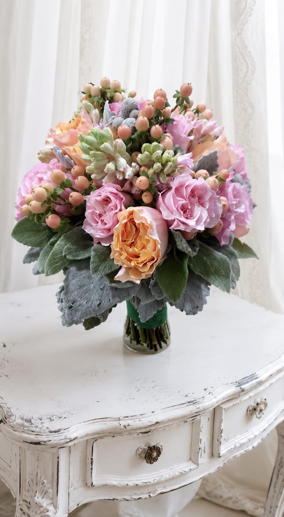 Pretty wedding bouquet - A soft cascade of pink and peach roses creates a delicate, feminine wedding bouquet that reads like vintage romance - perfect as a bridal centerpiece, anniversary gift, or spring wedding arrangement. The rounded, tightly clustered design immediately feels intimate and polished, the kind of bouquet that will make the person you're thinking of feel cherished and adored on a special day.  The arrangement pairs pale pink and peach roses with white highlights, muted gray-green foliage, and small berry-like accents for texture and a slightly weathered, romantic palette. Stems are finished with green floral tape and presented in a clear glass vase that keeps the focus on the dense, luxurious bloom mass while offering a timeless display for tabletops or a reception sweetheart table.  Designed to suit weddings, anniversaries, or thoughtful springtime surprises, this bouquet brings a calm, elegant presence to vows, milestone moments, or simply to say "you matter." Vase-ready and hand-tied for a neat silhouette, refresh water daily and trim stems every few days to help the roses stay fresh through the celebration.   If any design elements are of major importance to your order, please include them in the florist instructions at checkout or contact us to ensure availability.