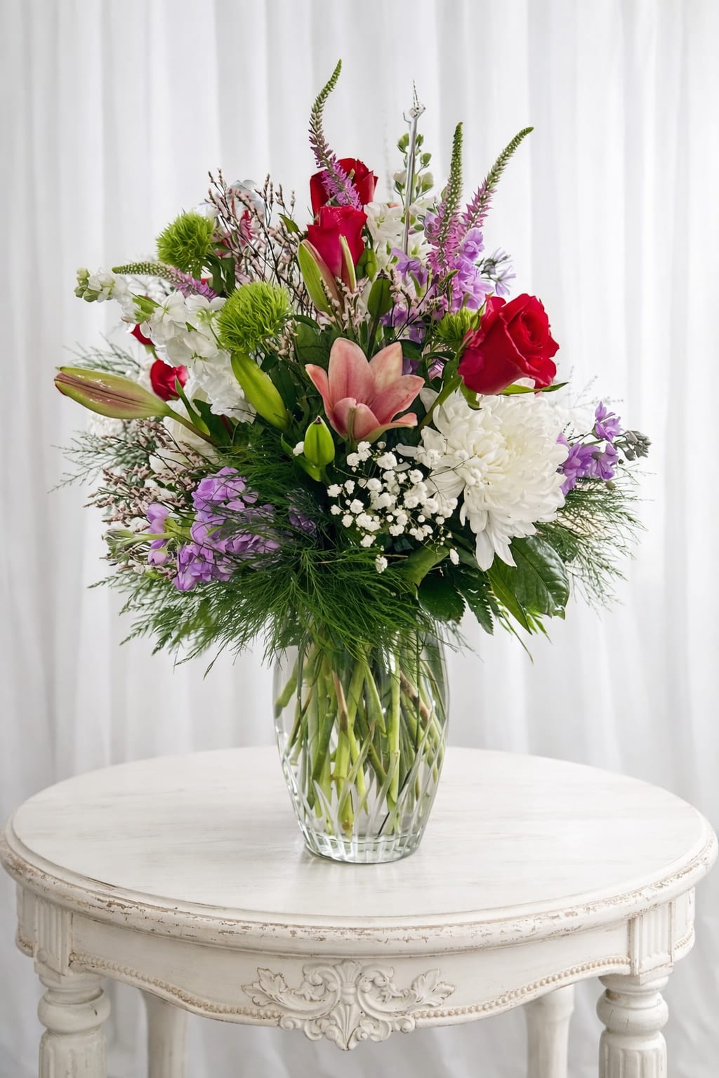 Good vibes - Tall and airy with a joyful mix of color, this flower arrangement is a celebratory mixed bouquet built to make someone feel truly seen. Perfect as a birthday bouquet, anniversary flowers, or a thoughtful sympathy arrangement, the composition pairs romantic red roses with pure white chrysanthemums, soft pink lilies, and vertical purple delphinium to create movement and optimism.  Layers of texture-velvety rose heads, trumpet-shaped lilies, fluffy mums, and delicate filler-rise from a clear glass vase that showcases the stems and keeps the design upright and abundant. The tall profile and long vertical spikes give the arrangement a graceful, uplifting silhouette that reads both fresh and elegant.  Send this when you want to lift their spirits or mark an important moment: she'll feel celebrated, comforted, or loved by the vibrant color story and thoughtful structure. Arrives ready to display in the included clear glass vase; change water every 2-3 days and trim stems to extend vase life.   If any design elements are of major importance to your order, please include them in the florist instructions at checkout or contact us to ensure availability.