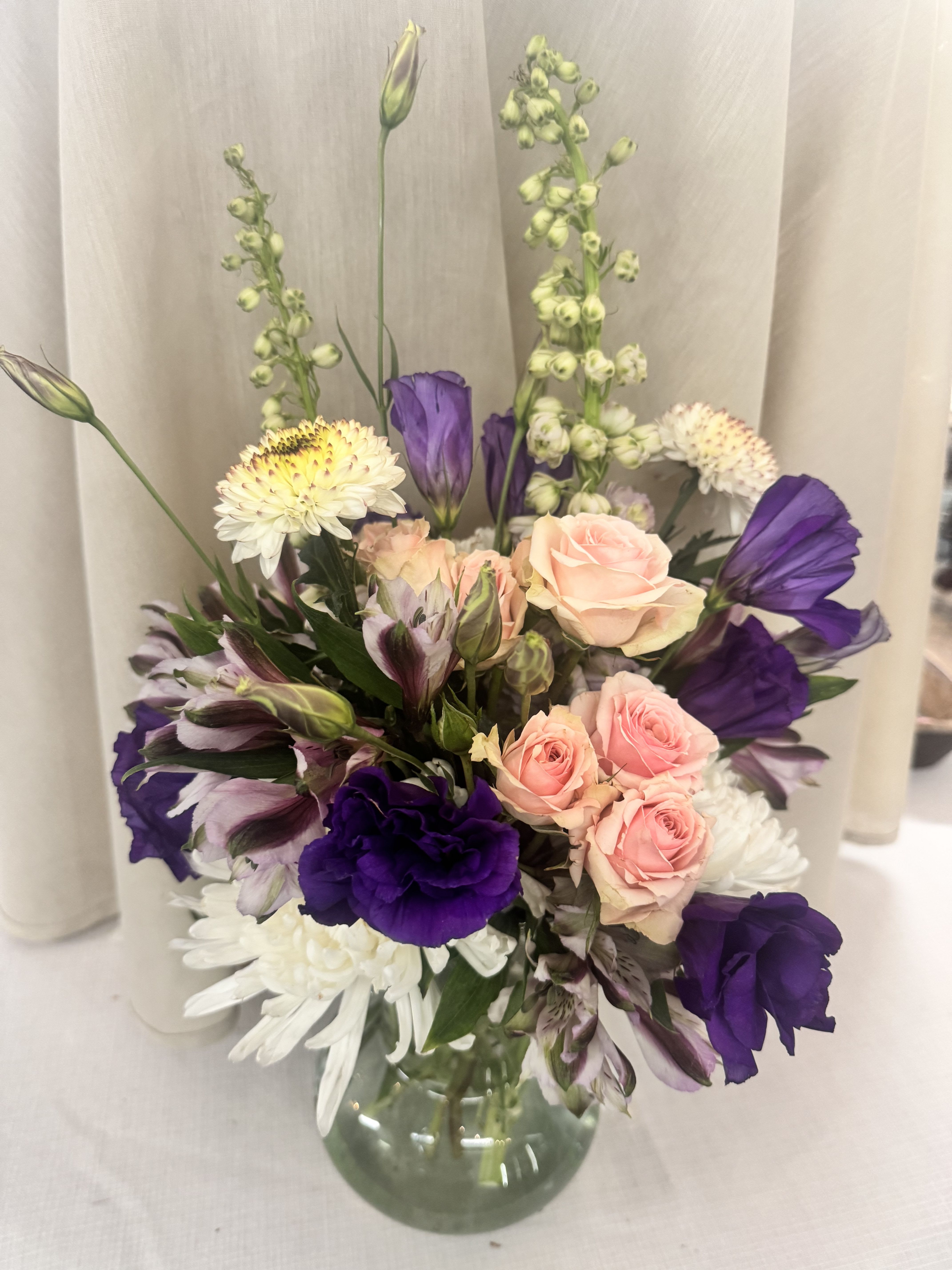 Purple Hayes - A lush mix of blush pink roses and saturated purple blooms gives this flower arrangement a romantic, garden-fresh look perfect for an anniversary bouquet, birthday flowers, or a bright thank-you gift. The vertical accents of tall green spikes add elegant height, so the recipient will feel celebrated by a design that's both full and airy.  This arrangement pairs white chrysanthemums and striped pink-and-white alstroemeria for layered texture and contrast, arranged in a clear glass vase for a clean, modern presentation. Colors: purple, pink, white, mixed; flowers include roses, alstroemeria, chrysanthemums, and complementary seasonal blooms. It's the kind of bouquet that feels thoughtfully arranged and lively-ideal for spring celebrations or anytime you want someone to feel truly seen.   If any design elements are of major importance to your order, please include them in the florist instructions at checkout or contact us to ensure availability.