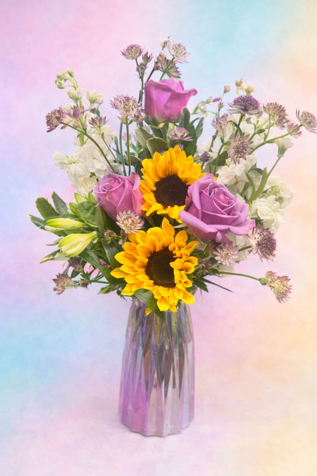 Just for you - A tall, airy bouquet that pairs cheerful sunflowers with soft lavender roses-perfect as a birthday bouquet, a thank-you gift, or a bright spring celebration arrangement. The mix of warm golden sunflowers and cool purple roses creates an immediate joyful contrast that will make the person you're thinking of feel noticed and uplifted.  This flower arrangement also includes white alstroemeria and small star-shaped filler blooms woven through fresh green foliage for texture and a garden-fresh look. Delivered in a clear glass vase that showcases the hand-arranged stems, the design reads both romantic and playful while staying simple and elegant.  Choose this for someone who loves a balance of bold color and delicate detail-the combination sends a message of warmth and thoughtfulness. Vase-ready and easy to display, refresh the water every 2-3 days and trim stems slightly to help keep the blooms lively.   If any design elements are of major importance to your order, please include them in the florist instructions at checkout or contact us to ensure availability.