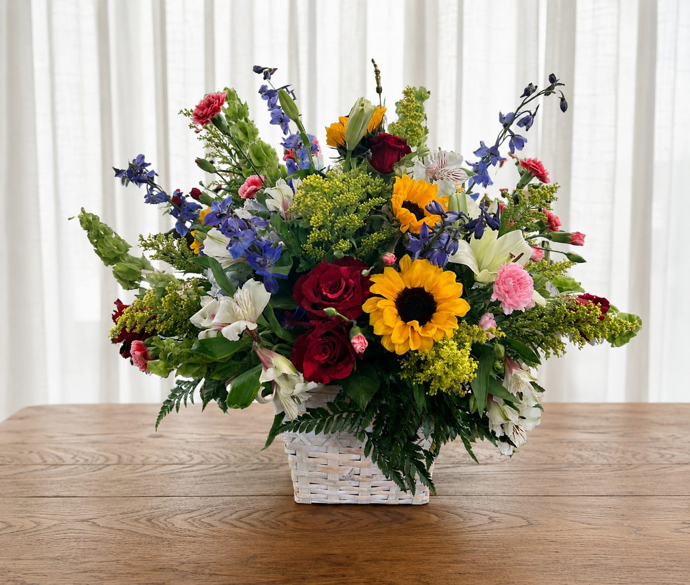 Basket of joy - A bright, garden-fresh centerpiece that reads like a celebration in bloom-this large mixed flower arrangement in a white woven basket is perfect as a birthday bouquet, a cheerful congratulations gift, or a warm everyday thank-you. The lively mix of vivid yellow sunflowers and deep red roses creates instant impact, while blue delphinium-like spikes and soft pink carnations add height and playful contrast so the recipient feels genuinely celebrated the moment they see it.  This airy, outward-spreading design also features white alstroemeria and complementary filler blooms for a layered, full presentation that looks hand-gathered from a sunny patch. Arranged in a classic white woven basket, the composition balances bold color and garden texture; it's generous in size and built to catch attention on a table, porch, or entryway. With its joyful palette and robust form, this basket makes a memorable gift that brightens any occasion.   If any design elements are of major importance to your order, please include them in the florist instructions at checkout or contact us to ensure availability.