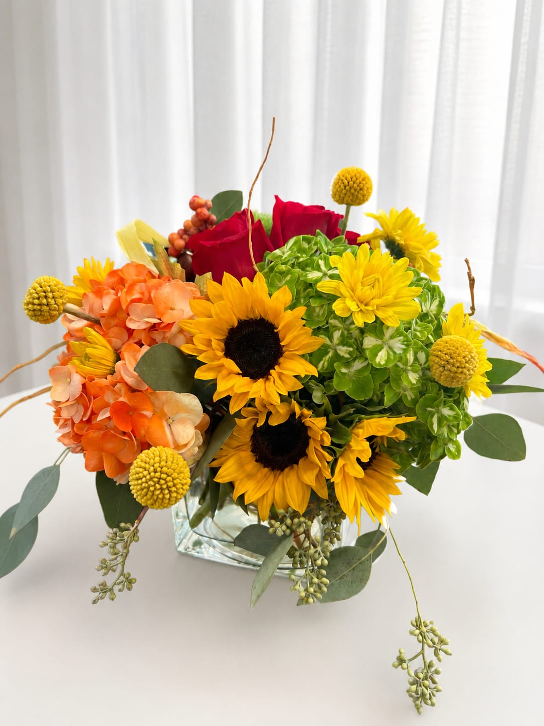Something special - Bright yellow sunflowers take center stage in this cheerful flower arrangement, designed as an everyday mixed bouquet perfect for birthdays, congratulations, or a bright thank-you gift. The combination of sunflowers and red roses with orange hydrangea creates a warm, celebratory palette, while green-and-white hydrangea and trailing eucalyptus-like foliage balance the look with fresh green accents-the kind of arrangement that makes the recipient feel genuinely seen and celebrated.  This compact arrangement is gathered in a clear glass vase that showcases stems and adds simple, modern polish. Textural pops come from round yellow pom chrysanthemums and clusters of hydrangea, giving layered volume and movement without feeling fussy. The mix of large focal blooms and smaller pom tones creates a lively visual contrast and a pleasant, garden-fresh scent profile.  Perfect for sending to a friend, coworker, or family member who could use an energetic pick-me-up, this bouquet reads as effortless joy-bright, warm, and thoughtfully arranged. Vase-ready and arranged for immediate display, it's an easy, confidence-building choice when you want to make someone's day.   If any design elements are of major importance to your order, please include them in the florist instructions at checkout or contact us to ensure availability.