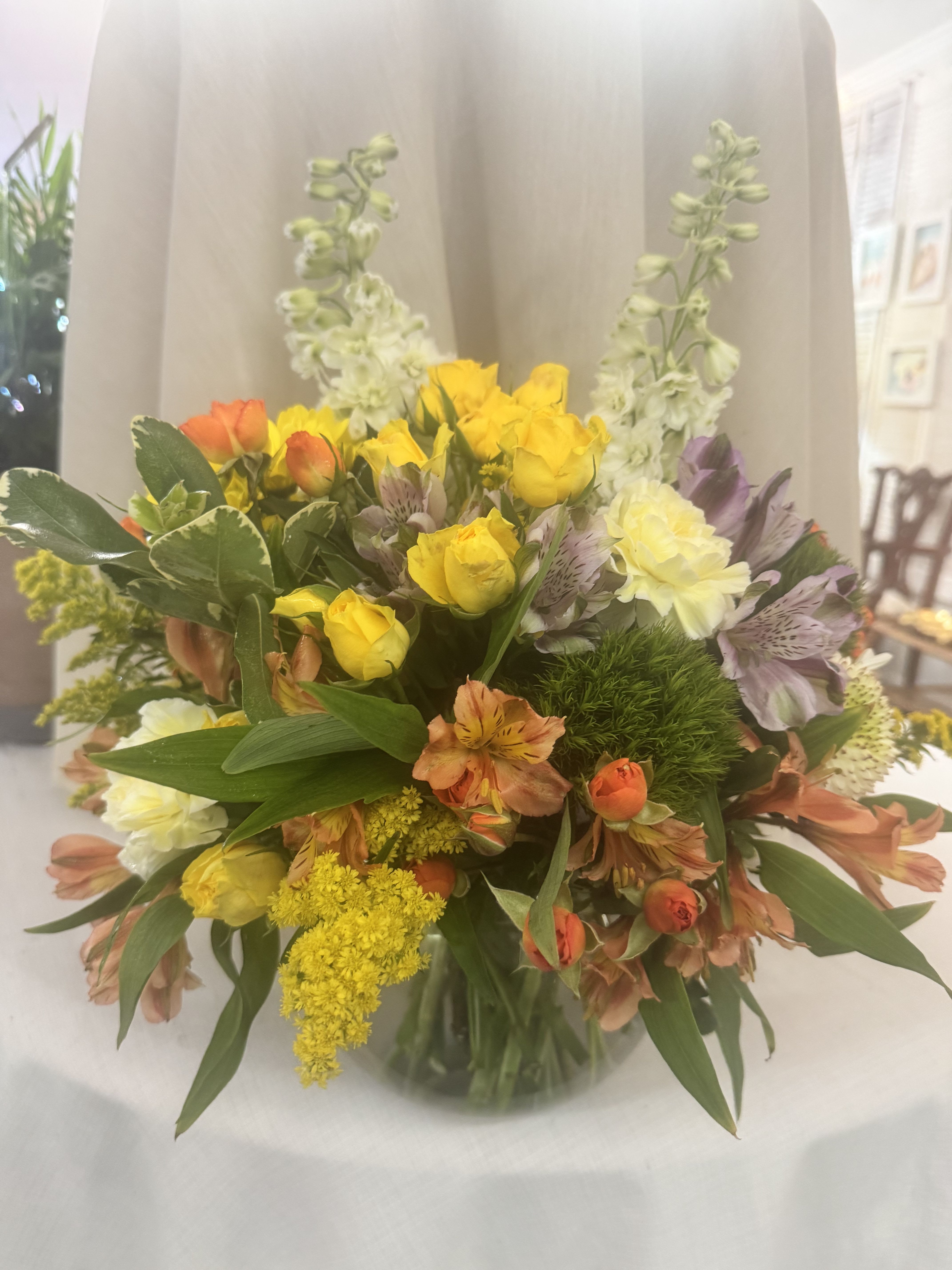 Blooms for you - Bright yellow roses take center stage in this everyday celebratory flower arrangement, a cheerful bouquet perfect for birthdays, thank-you gifts, or brightening a spring table. The mix of sunny yellow and warm orange with soft lavender accents creates a lively, garden-style impression that makes the person you're thinking of feel genuinely celebrated.  Arranged in a clear glass vase, the design pairs yellow roses with orange alstroemeria, pale cream chrysanthemums and other soft blooms for a full, rounded silhouette. Tall pale green-white spikes rise behind the main cluster to add vertical interest and a light, airy frame. Colors include yellow, orange, white and purple; the vase gives it a polished, ready-to-display finish.  This is the kind of bouquet that suits neighbors, coworkers, moms or friends-anyone who'd appreciate a bright, optimistic surprise. Standard care: keep stems in fresh water, trim a half-inch before placing and refresh water every other day to help blooms last.   If any design elements are of major importance to your order, please include them in the florist instructions at checkout or contact us to ensure availability.