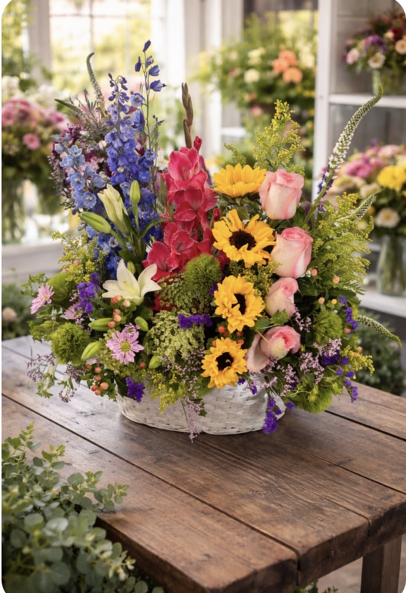 English garden - A cheerful, garden-fresh display that reads like a sunlit cottage border - this English garden flower arrangement is a large mixed-color bouquet perfect for birthdays, congratulations, or any spring celebration. The tall vertical accents and rounded blooms create a lively, layered silhouette that will make the person you're thinking of feel genuinely celebrated and seen.  Rich yellow sunflowers set a sunny tone alongside pink roses, blue delphinium, red snapdragons, white lilies, daffodils, alstroemeria and textured filler for an airy, abundant look. The flowers come arranged in a white wicker basket, giving the design a relaxed, hand-picked vibe with pleasing height and fullness. Colors include pink, purple, yellow and mixed bright tones for a festive, joyful impression.  Ideal for surprising a friend, brightening a workplace, or marking a special milestone, this arrangement delivers an immediate mood lift and feels effortless to display. Care is simple: keep water fresh and trim stems every few days to extend vase life, and choose delivery during spring or anytime you want to send warm, colorful cheer.   If any design elements are of major importance to your order, please include them in the florist instructions at checkout or contact us to ensure availability.