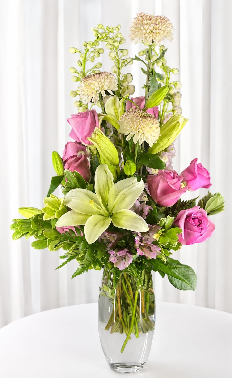 Pretty pink - Soft pink roses and pale lilies rise above a cloud of creamy chrysanthemums in a tall, layered birthday bouquet that reads both romantic and joyful. This flower arrangement for birthdays, anniversaries, or a cheerful congratulations delivers a fresh, airy impression-perfect for the person you want to make feel cherished and celebrated.  Arranged in a clear glass vase, the upright design showcases stems through the glass and keeps the look clean and modern. The composition pairs pink roses, ivory lilies, cream chrysanthemums and mixed-color alstroemeria with bright green buds for contrast, creating pleasing height and texture that's pretty from every angle.  Send this piece when you want her to feel genuinely seen-whether marking an anniversary, celebrating a milestone, or simply brightening someone's day. The tall vase gives it presence on a dining table or entry console; typical care is fresh water and a cool spot away from direct sunlight to help the blossoms last.   If any design elements are of major importance to your order, please include them in the florist instructions at checkout or contact us to ensure availability.