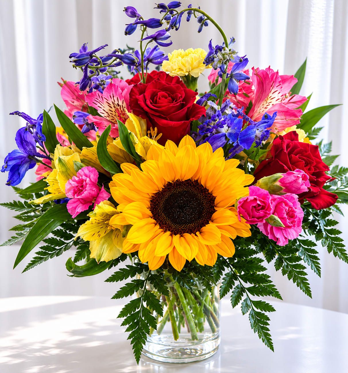 Sunflower delight - A bold golden sunflower takes center stage in this cheerful flower arrangement-perfect as a birthday bouquet, a congratulations gift, or a general pick-me-up. The mix of sunflowers and classic roses creates an immediate sunny impression while the pink alstroemeria and carnations add soft, affectionate notes; together they say "you're celebrated" to the person you're thinking of.  This mixed-color arrangement pairs a prominent yellow sunflower with red roses, pink alstroemeria, pink carnations and small blue‑purple accent blossoms, all gathered in a clear glass vase. Broad, fern-like foliage spills around the base for a full, layered silhouette and lively texture that reads beautifully across a table or entryway.  Give this to someone who could use an instant lift-the bright contrasts and joyful colors make her feel noticed and celebrated. Arrives ready to display in its glass vase; simple fresh-water care will keep the blooms vibrant for days.   If any design elements are of major importance to your order, please include them in the florist instructions at checkout or contact us to ensure availability.