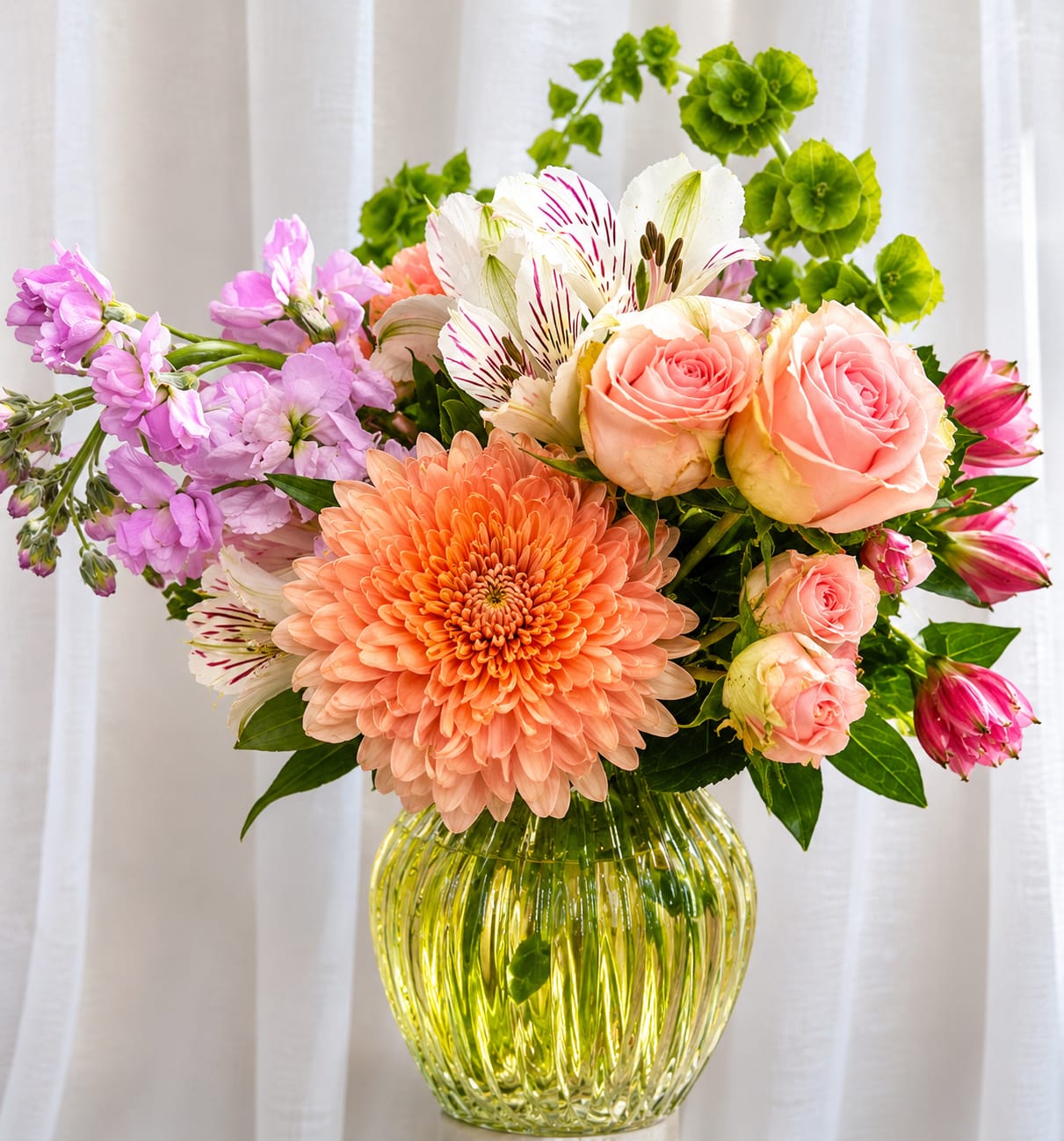 Blushing Garden Grace - A joyful mix of peach-orange chrysanthemum and blush roses makes this flower arrangement an uplifting choice for a birthday bouquet or spring celebration. Blushing Garden Grace sends a warm, celebratory message-perfect for the person you're thinking of who loves a romantic palette with a playful twist-so she'll feel noticed and brightened the moment these flowers arrive.  The arrangement pairs a large peach-orange chrysanthemum with soft blush roses, purple stock, white alstroemeria marked in magenta, and lively bright green accent blooms that add height and contrast. Tucked into a clear ribbed yellow glass vase, the composition reads full and fresh: pastel petals, textured stock, and crisp green accents combine for a balanced, modern look that suits a table centerpiece or an everyday gift.  Ideal for birthdays, springtime thank-yous, or cheering someone up, this arrangement arrives ready to display in its decorative vase. For best longevity, place it in a cool spot away from direct sun and change the water every other day to keep blooms looking their freshest.    If any design elements are of major importance to your order, please include them in the florist instructions at checkout or contact us to ensure availability.