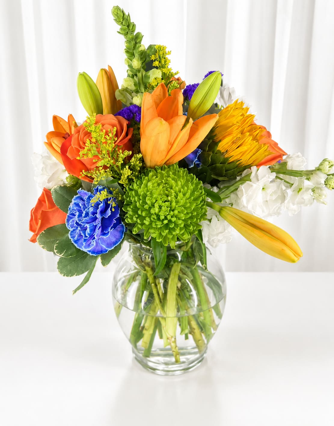 Sunlit Celebration - Sunlit Celebration greets the recipient with a burst of warm color-an everyday bouquet perfect for birthdays, congratulations, or spring celebrations. This flower arrangement captures a cheerful, energetic mood with orange and yellow blooms balanced by vivid green accents and bright touches of white and blue, the kind of gift that makes someone feel noticed and genuinely celebrated.  The mix pairs tulip buds, ruffled carnations, garden roses, snapdragons, and a standout green chrysanthemum, finished with small filler flowers for texture. Arranged in a clear glass vase, the composition is full and layered so each stem shows off its shape and color; tactile ruffles and smooth tulip cups create lively contrast. The palette reads sunny and fresh-ideal for sending optimism or marking a joyful milestone.  Designed for easy enjoyment, the vase-ready format arrives ready to display on a table or desk and suits recipients who appreciate bright, seasonal blooms. To keep the bouquet looking its best, refresh the water every few days and trim stems at an angle-small care actions that help this vibrant arrangement continue to brighten their day. If any design elements are of major importance to your order, please include them in the florist instructions at checkout or contact us to ensure availability.  
