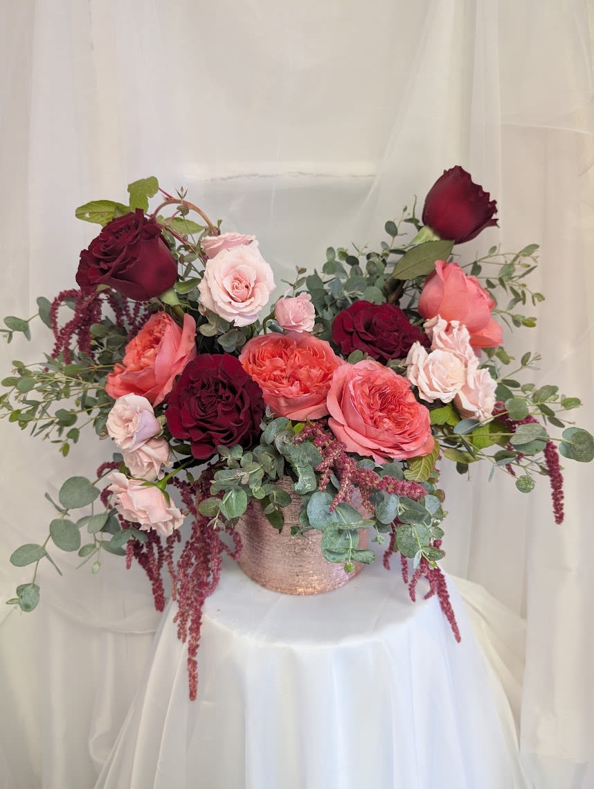 A Shirley Temple  - A modern one-sided arrangement with a combination of Garden Roses, Spray Roses, Hanging Amaranthus accents and greenery. Arranged beautifully in a rose gold vase.  