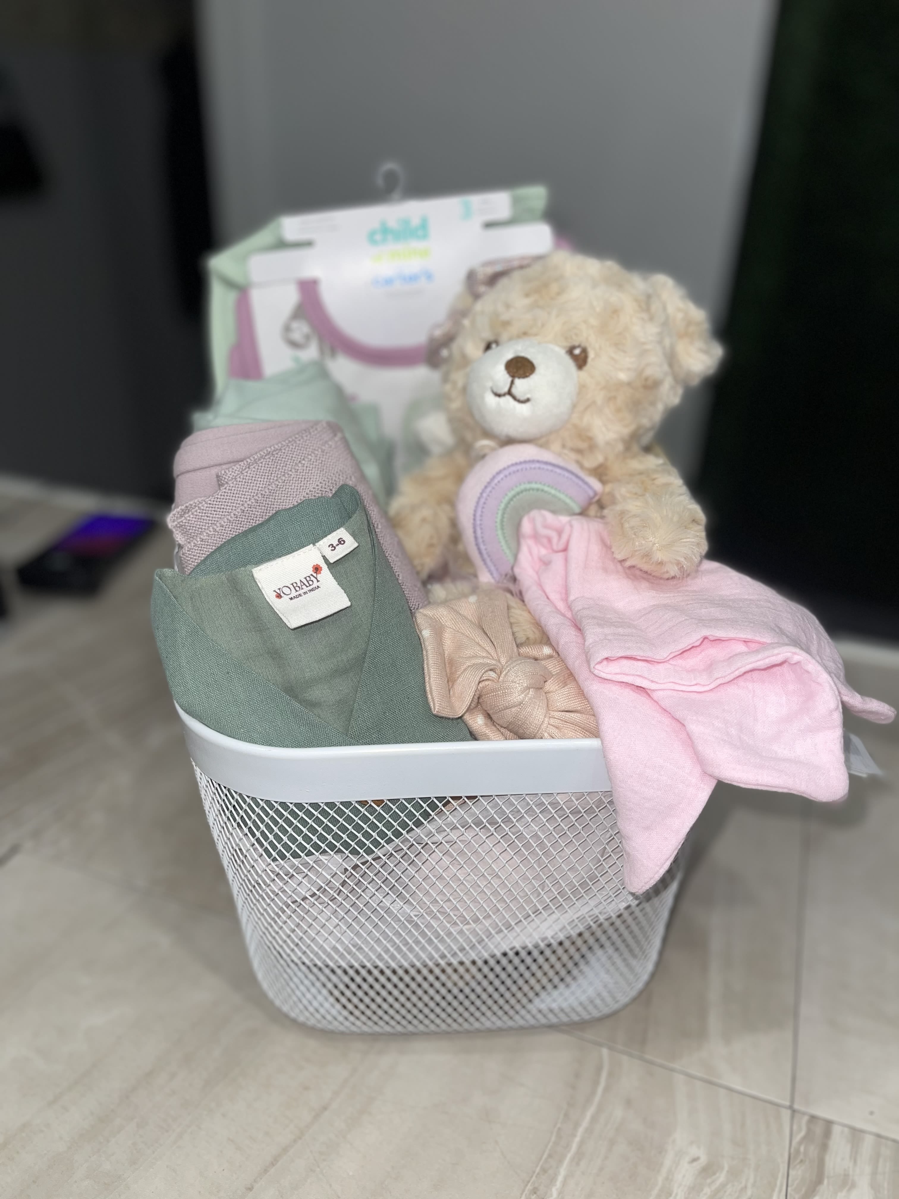 Hello Baby Girl Basket - Soft pastels and cuddly textures make this Hello Baby Girl Basket the kind of newborn gift that instantly comforts and delights. Perfect as a new baby or baby shower present, the baby gift basket reads like a thoughtful welcome-home surprise for the little girl you're celebrating - she'll be surrounded by soft blankets, plush friends, and sweet essentials that set a gentle, nursery-ready tone.  Arranged upright in a white wire or woven basket, this set includes a plush teddy or doll, folded baby clothing and blankets in pink, mint, lavender, peach and cream, plus curated extras like a scented candle jar, packaged accessories, baby bottles, and hair bows. The layered presentation and mixed textures - soft knits, cotton garments, and wicker or mesh basket - create a tidy, gift-ready look that's easy to place in the nursery.  This basket is ideal for the new mom who could use cozy essentials and a little pampering; it's the kind of present that says "congratulations" while helping baby feel snug and loved. Choose size and packaging options at checkout   Basket items may vary due to availability.*