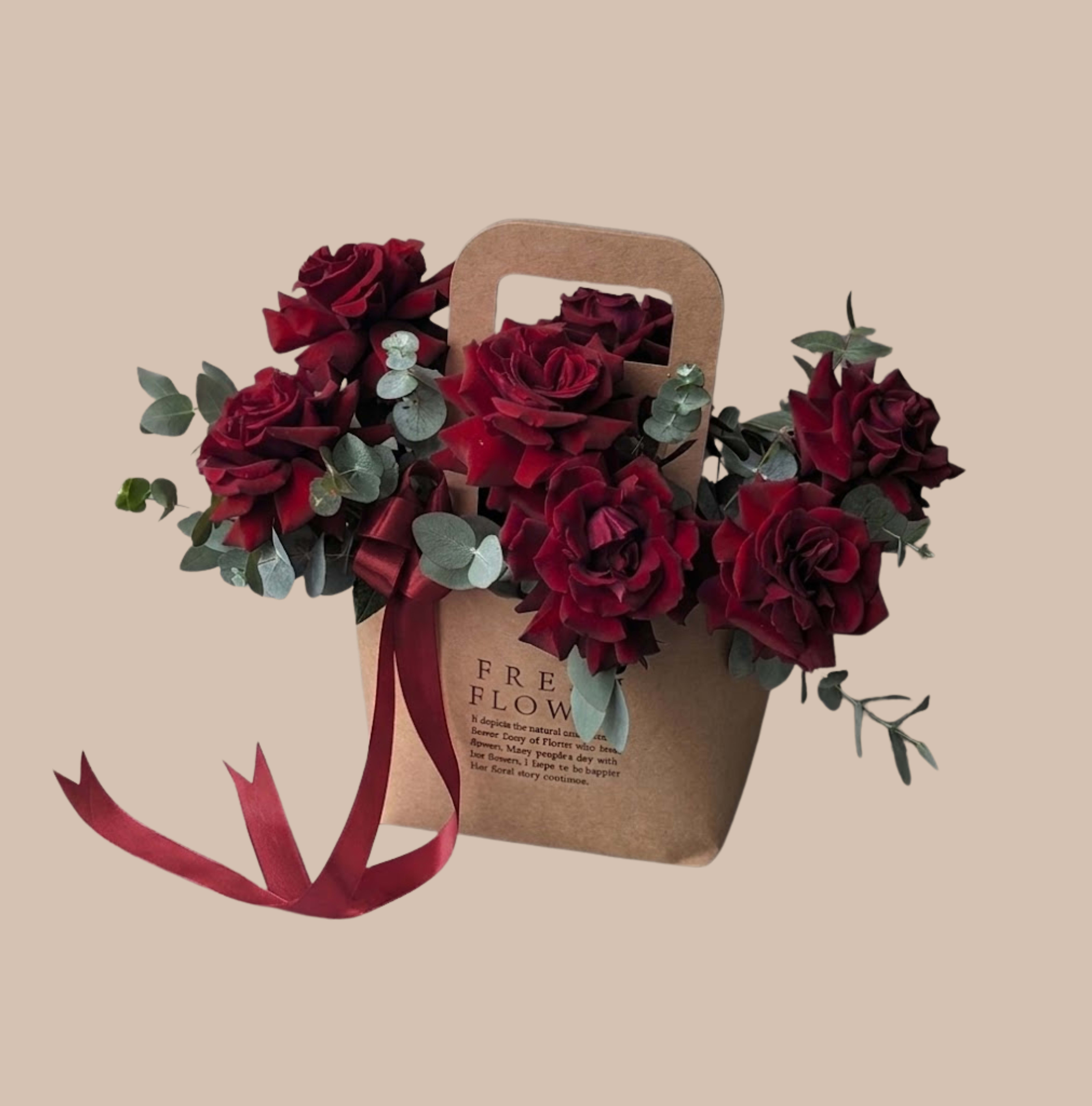 Kraft &amp; Crimson Carrier - Deep burgundy roses tucked into a kraft paper gift bag create an intimate, gift-ready statement perfect for an anniversary bouquet, romantic surprise, or a heartfelt thank-you. The rich red and crimson tones read as classic romance while the compact carrier and satin ribbon make the presentation feel deliberate - the person you're sending it to will feel genuinely celebrated the moment they see it.  Clusters of velvety roses are balanced by soft eucalyptus sprigs that add a muted gray-green contrast and gentle texture. The arrangement sits inside a cutout-handle kraft paper gift bag, tied with a matching burgundy satin ribbon for a coordinated, polished look that's easy to carry and display. Ideal for gifting straight from the shop, this piece arrives ready to present and brightens tabletops, desks, or bedside stands with moody, elegant color.