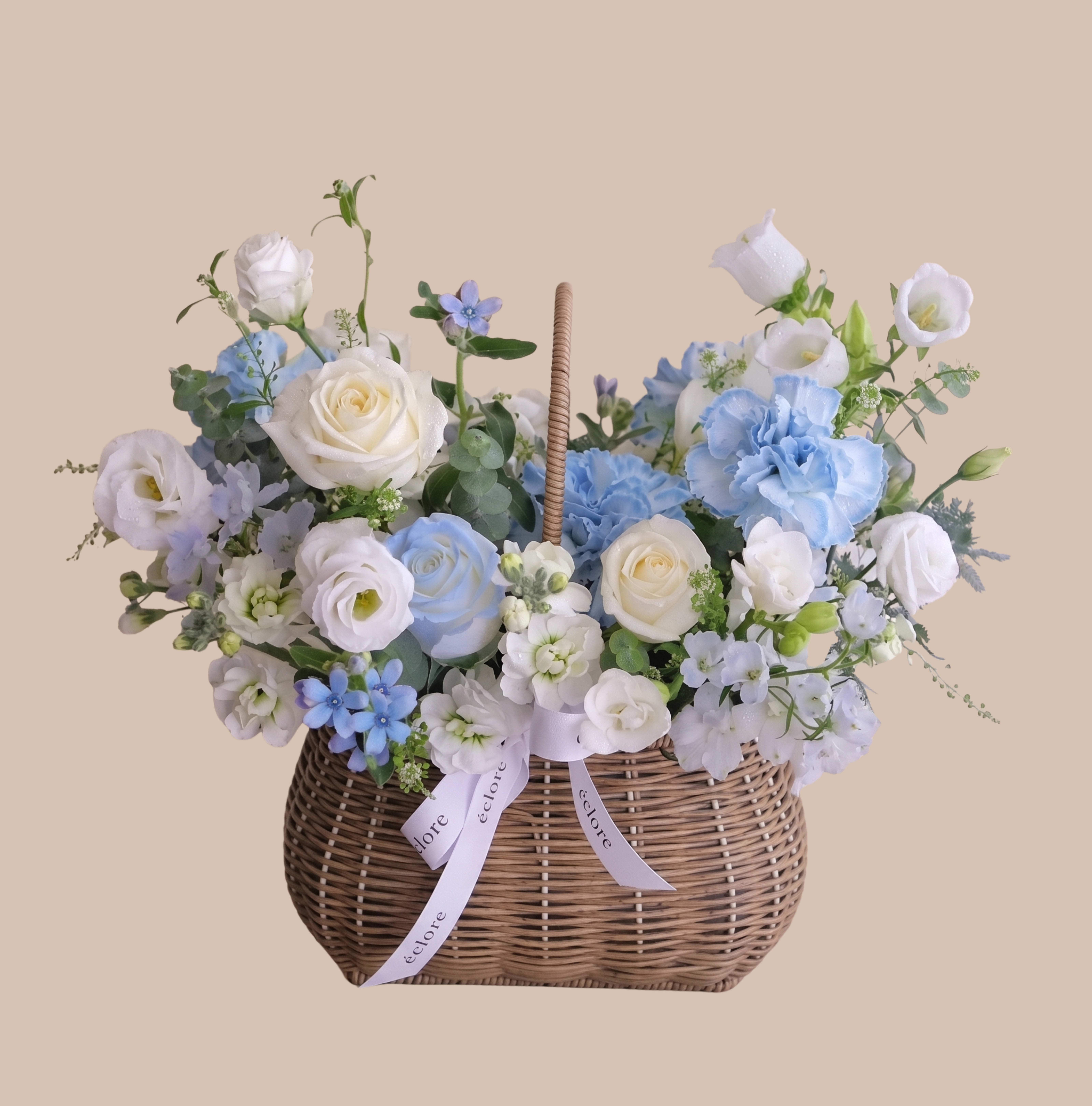 Blue Baby &amp; Basket - Soft blue and white blooms gathered in a woven wicker basket create a gentle, garden-fresh welcome for a new baby. This baby boy basket arrangement is a thoughtful new-baby gift or baby shower centerpiece that reads as calm and joyful - the kind of floral surprise that makes the new parents feel truly celebrated.  The rounded, abundant composition pairs white roses and lisianthus with pale blue delphinium, stock, and carnations, plus small accent flowers and greenery for airy texture and soft contrast. Arranged compactly in a wicker basket with a pale ribbon tied at the handle, the display has a homey, hand-tied look that sits neatly on a nursery dresser or side table and delivers immediate visual impact without a vase.  Ideal for welcoming a newborn, saying congratulations, or sending a gentle "thinking of you," this arrangement arrives ready to display. For best longevity, keep blooms in a cool spot away from direct sunlight and refresh any visible water in the basket as needed.