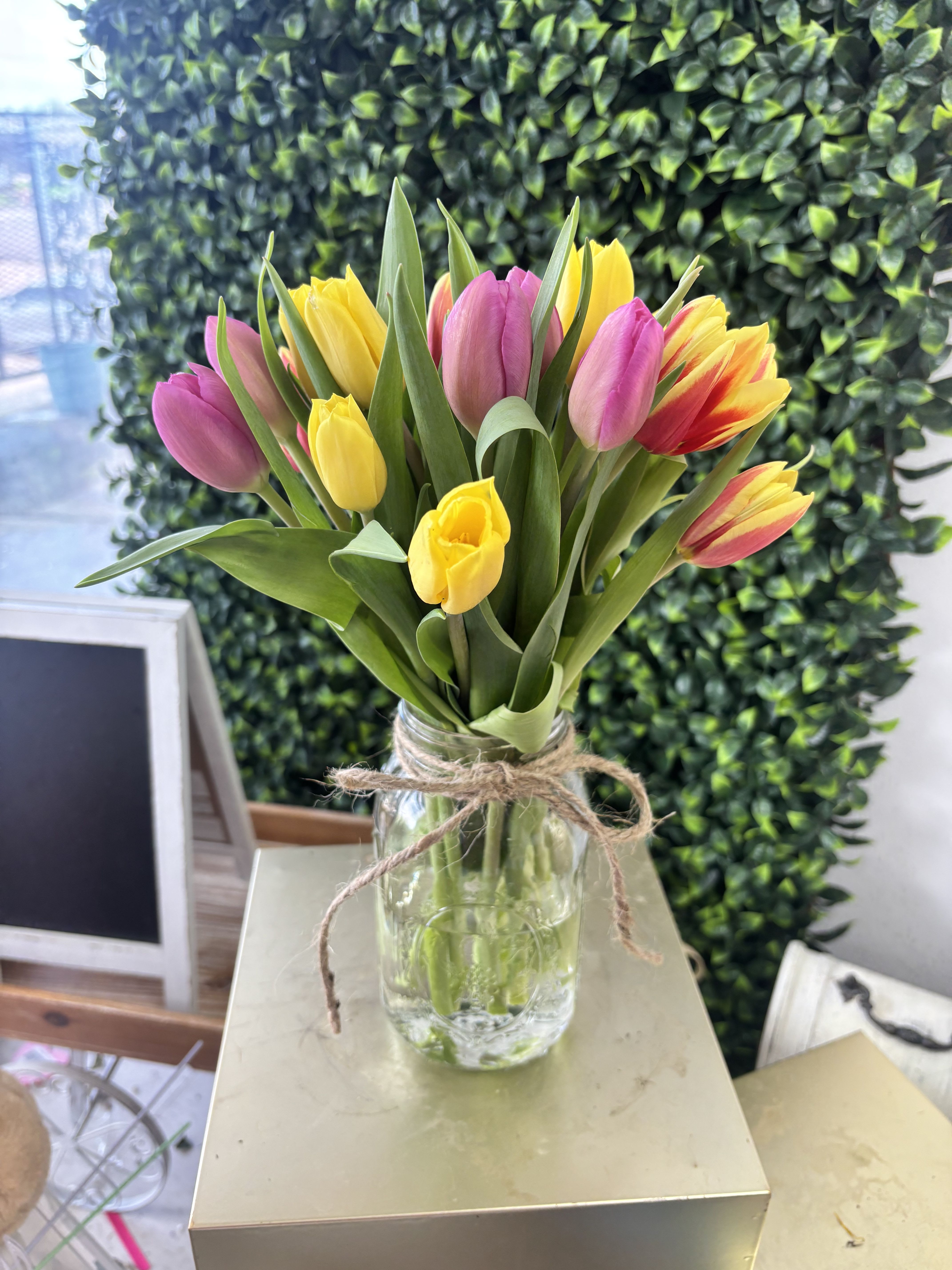 Tulip Garden  - A lively cluster of pink, yellow, and mixed-color tulips creates an instant spring moment - the perfect birthday bouquet or cheerful everyday gift. This tulip arrangement for seasonal celebrations feels bright and effortless, the upright blooms and fresh green leaves giving the person you're thinking of a feeling of sunlit optimism.  Gathered in a clear glass jar tied with a simple twine bow, the design reads casual and homey while letting the tulips take center stage. Long green foliage frames the vibrant petals so the display looks full without being fussy; proportions are ideal for a tabletop, mantel, or kitchen counter. It's a thoughtful way to say "happy day" or "thinking of you" for anyone who enjoys a fresh, seasonal touch.