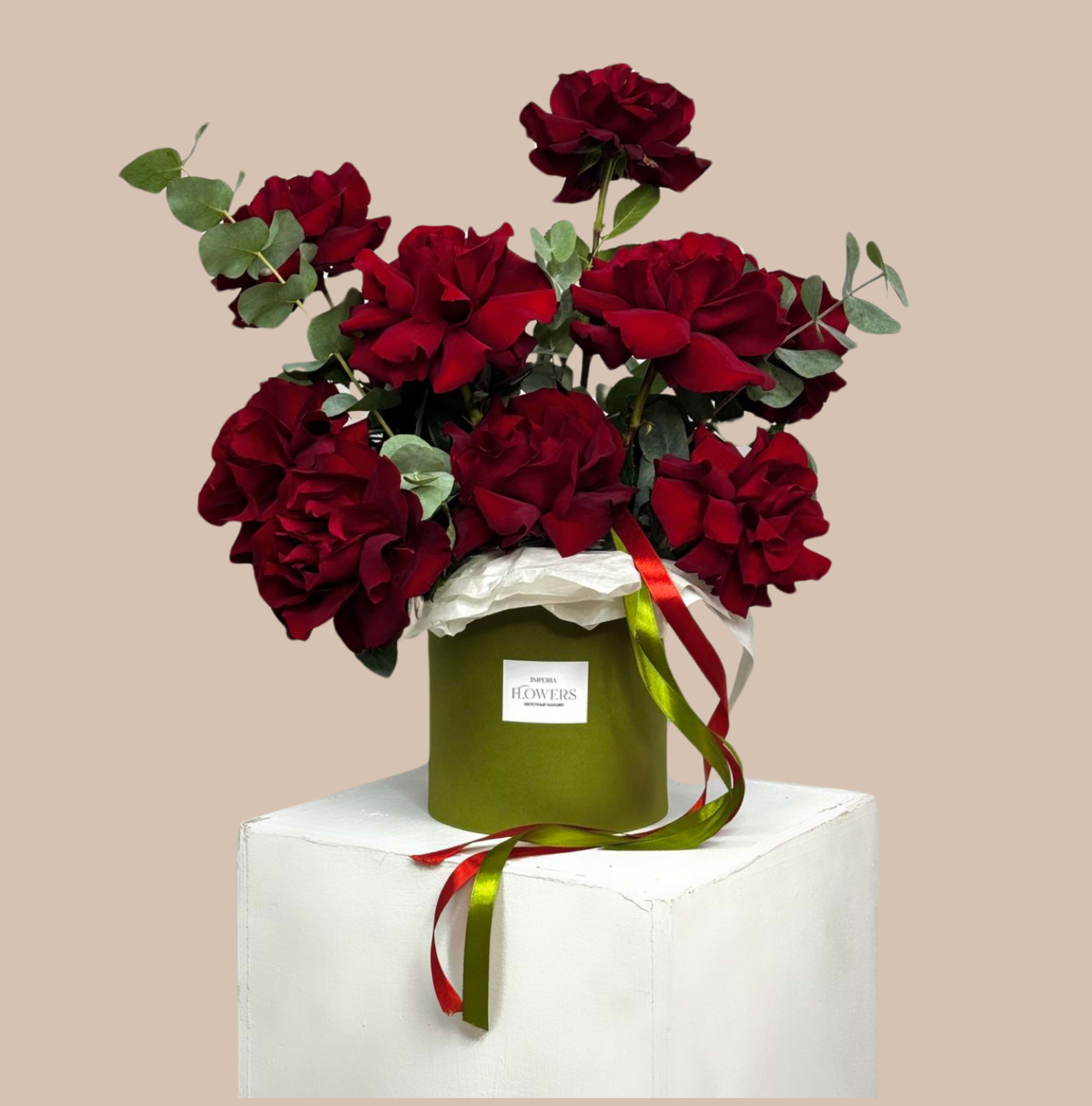Moss &amp; Merlot - A lush cluster of deep red roses rises from a round green hat box, the perfect romantic bouquet for a Valentine's Day surprise, anniversary roses, or any occasion when you want someone to feel truly adored. The velvety, full blooms read classic and intimate while the eucalyptus-like foliage adds a soft gray-green contrast, making the recipient notice both color and texture the moment they see it.  Arranged in a green hat box with white tissue paper at the top, this floral gift is finished with long red and green ribbons and a branded label for a polished presentation. The compact round shape makes it easy to place on a table or desk; the dense arrangement keeps the roses visually impressive without overwhelming small spaces.  Choose this arrangement when you want to make someone feel seen and cherished-whether marking a milestone anniversary or sending a thoughtful celebration gift. Designed for immediate display, enjoy simple care: keep the box in a cool spot away from direct sun and replace water as needed to prolong bloom life.