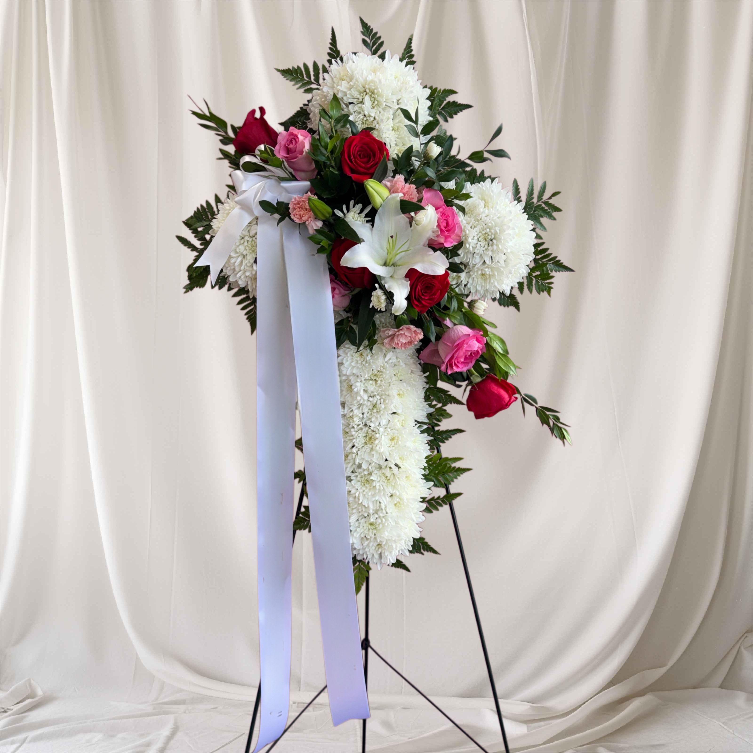 Cross of lightt - A tall standing cross of white chrysanthemums anchored with clusters of red and pink roses creates an immediate, reverent presence-perfect as a sympathy arrangement or memorial tribute. The white lily bloom near the center lends a quiet focal point while touches of green foliage and soft pink carnations add gentle texture, so the recipient feels honored and remembered.  This formal spray is finished with a long white ribbon and presented on a black metal easel for dignified display at a service or graveside. The dense white chrysanthemum base, mixed rose accents, and single prominent lily combine classic shapes and comforting colors to convey sympathy and respect. Order with confidence knowing this design is crafted for ceremony and remembrance; care is simple-keep in a cool, sheltered location and replace water for any vase components if removed from the easel.