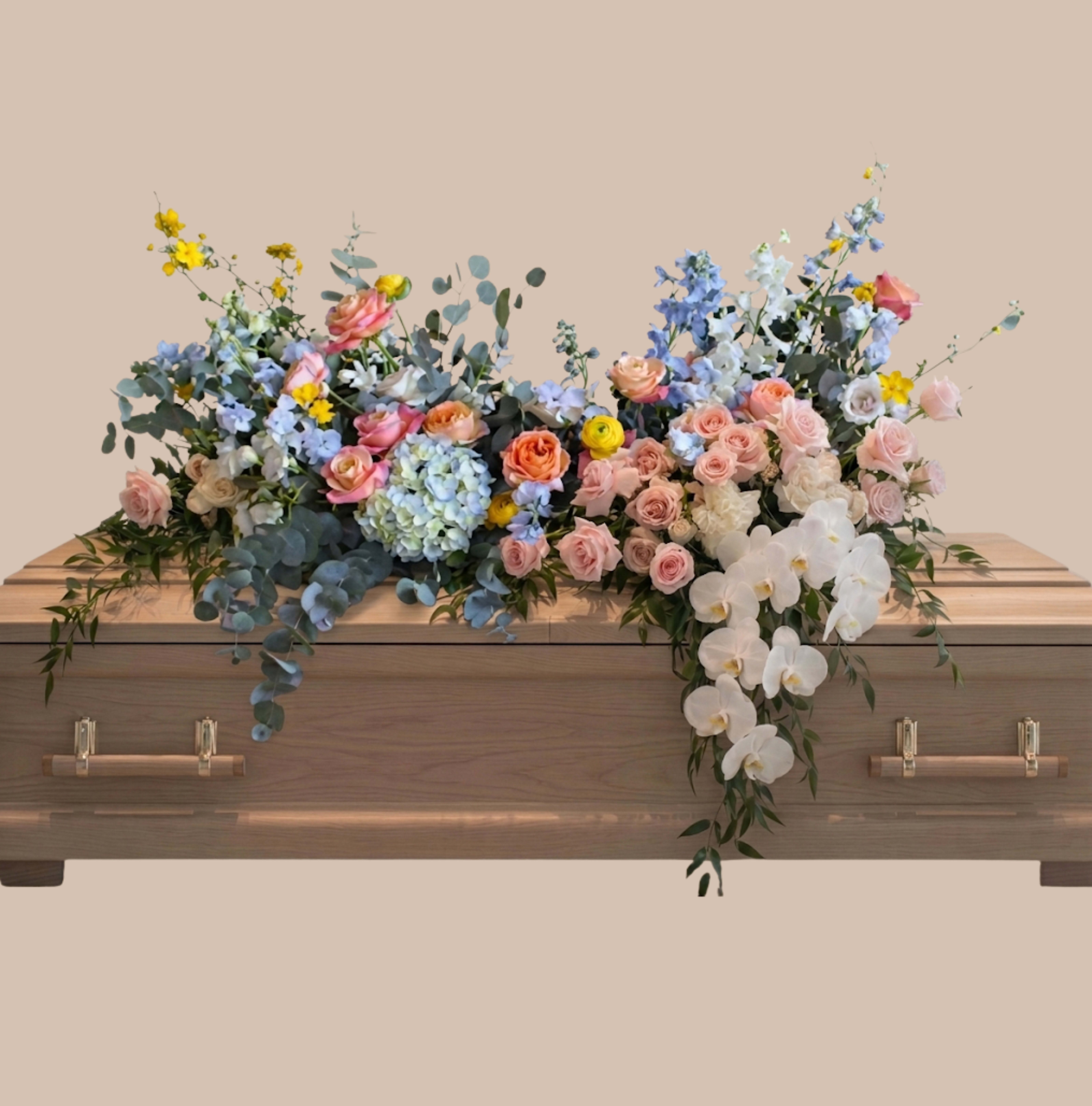 Pastel Clouds Spray - Soft pink roses, pale blue hydrangeas, and graceful white orchids are arranged in a wide, asymmetrical casket spray designed to convey tenderness and calm-an elegant funeral or memorial tribute that soothes while honoring. The pastel palette, brightened with airy blue delphinium and small yellow accents, creates a soft, cloudlike impression that will help the person you're remembering feel seen and cherished.  This spray is arranged across the top of a wooden casket, spilling outward in a lush, layered composition accented with eucalyptus for texture and gentle greenery. The mix of roses, hydrangeas, orchids, delphinium, and other seasonal blooms offers both full-bodied presence and delicate details; the overall scale and formal styling make it appropriate for traditional services and intimate gatherings alike.  Choose this piece when you want to express sympathy with quiet beauty-a meaningful way to say you're thinking of the family and honoring a life. For sizing, placement, or delivery questions specific to casket arrangements, please contact us so we can tailor the spray to the service and ensure it arrives fresh and respectfully arranged.
