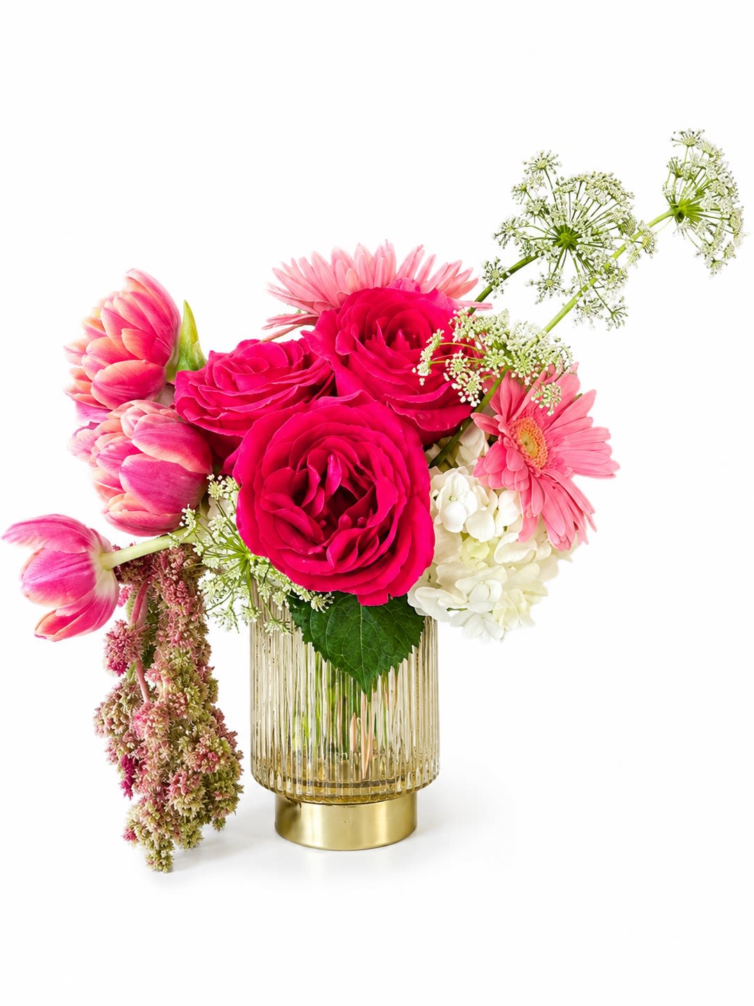 Pink Martini - Hot-pink roses take center stage in this lively flower arrangement, making it the perfect birthday bouquet or Valentine's Day surprise for someone who loves bold, romantic color. The mix of pink tulips and a single pink gerbera daisy around a cloud of white hydrangea reads both joyful and elegant, so the person you're thinking of will feel genuinely celebrated the moment these blooms arrive.  The composition is airy and slightly asymmetrical, with delicate white filler flowers and branching stems reaching upward and to the right for a fresh, springtime silhouette. Colors are bright pinks balanced with crisp white and hints of green and sunny yellow; flowers include roses, tulips, gerbera daisy, hydrangea and seasonal accents. The arrangement sits in a ribbed glass vase finished with a gold-toned base, giving a polished, modern contrast to the soft petals and making it ready to display on a table or mantle.  Ideal for anniversaries, congratulations, or an effortless "thinking of you," this design reads celebratory without being overdone. Size and care: flowers are arranged for immediate display in the included glass vase - trim stems and refresh water every few days to keep blooms perky.