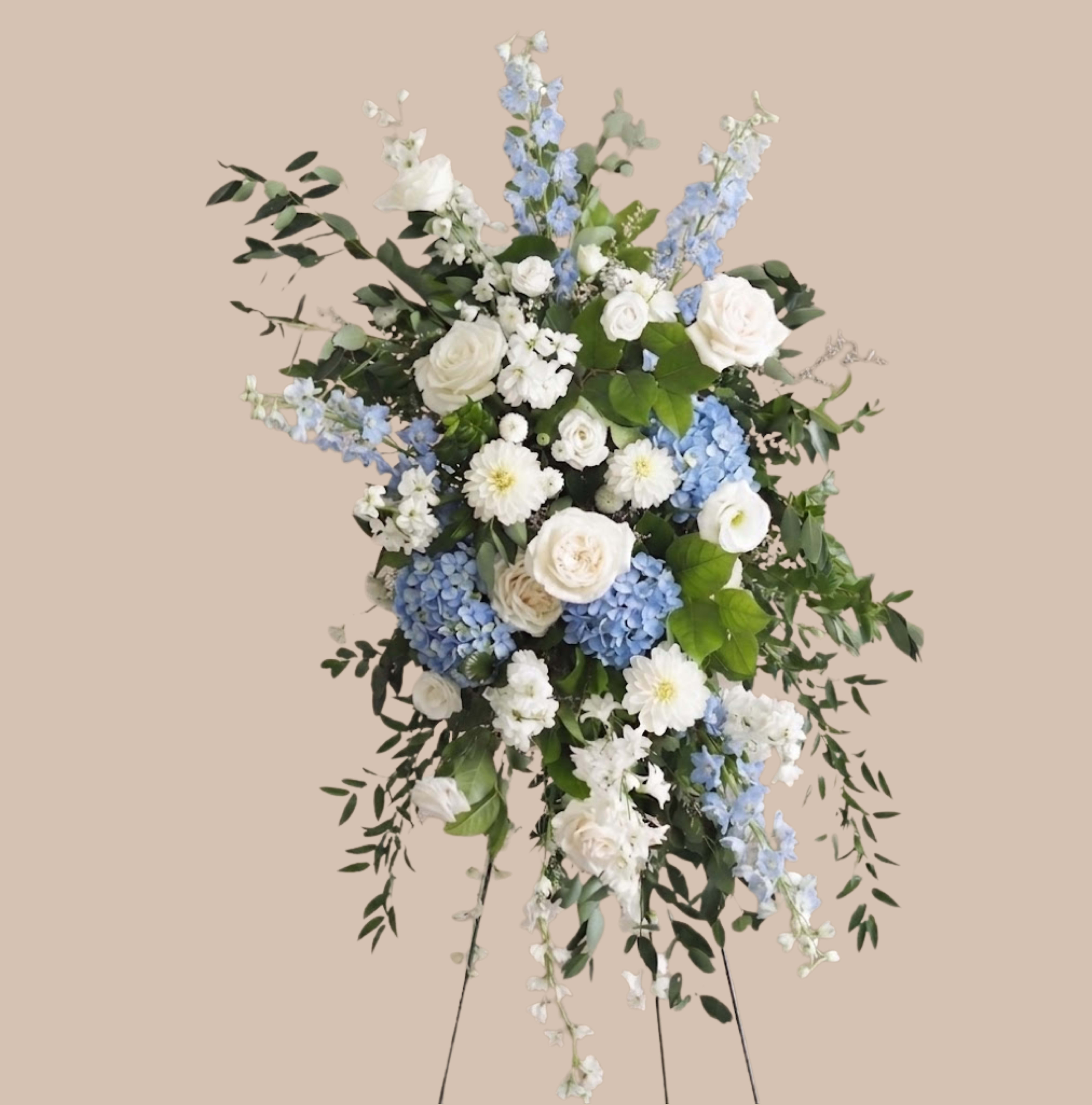 Pillars of Peace Easel - A serene, upright sympathy easel that speaks of quiet comfort and graceful remembrance-this standing spray is designed for funerals and memorial services. The Pillars of Peace Easel pairs classic white roses and garden roses with soft daisy-like blooms to create a luminous, comforting face, while cool blue hydrangeas and pale delphinium spires add a gentle, contemplative contrast. Presented on a standing easel, its formal silhouette reads as a thoughtful tribute that will make the person you're honoring feel remembered and respected.  Layered greenery and lightly cascading foliage give the arrangement an airy, vertical flow; ranunculus, stock, and small daisies contribute delicate texture among the larger blooms. The color palette of white, green, and blue keeps the impression calm and refined, suitable for traditional services or a private memorial. Sized and arranged for visibility when displayed on an easel, this spray is ready to place at a graveside service or church front; blooms are selected and conditioned for freshness, and local delivery/setup can be arranged to ensure it arrives properly presented.