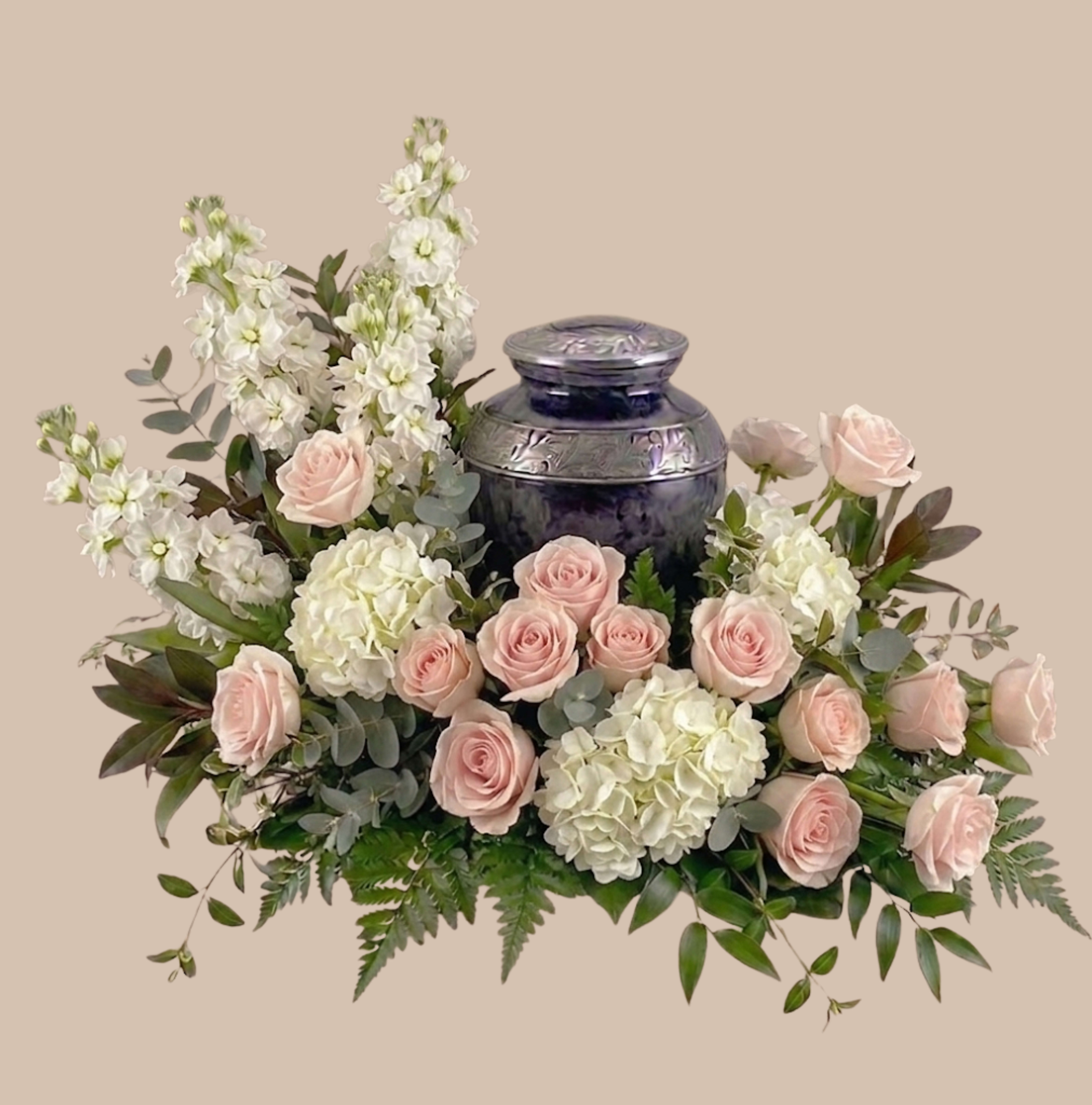 Blush Elegance Tribute - A serene bed of pale pink roses, white hydrangeas, and tall white stock rests in a dark decorative urn, creating a Blush Elegance Tribute designed for sympathy and funeral settings. This formal flower arrangement reads as a graceful, comforting centerpiece-the soft blush and cool white palette paired with layered greenery offers a quiet, consoling presence the person you're honoring will look dignified beside.  Carefully composed as a low, wide display rather than a handheld bouquet, the design also includes garden roses, spray roses, ranunculus and tulips arranged for full symmetry and gentle texture. The dark urn grounds the piece and heightens the pale florals' tones; the taller stock provides vertical interest while the hydrangeas and mixed blooms create lush volume. Ideal for memorial tables, viewing rooms, or as a thoughtful home tribute, the arrangement arrives ready to display-no special care beyond fresh water is required to keep it looking composed through services.