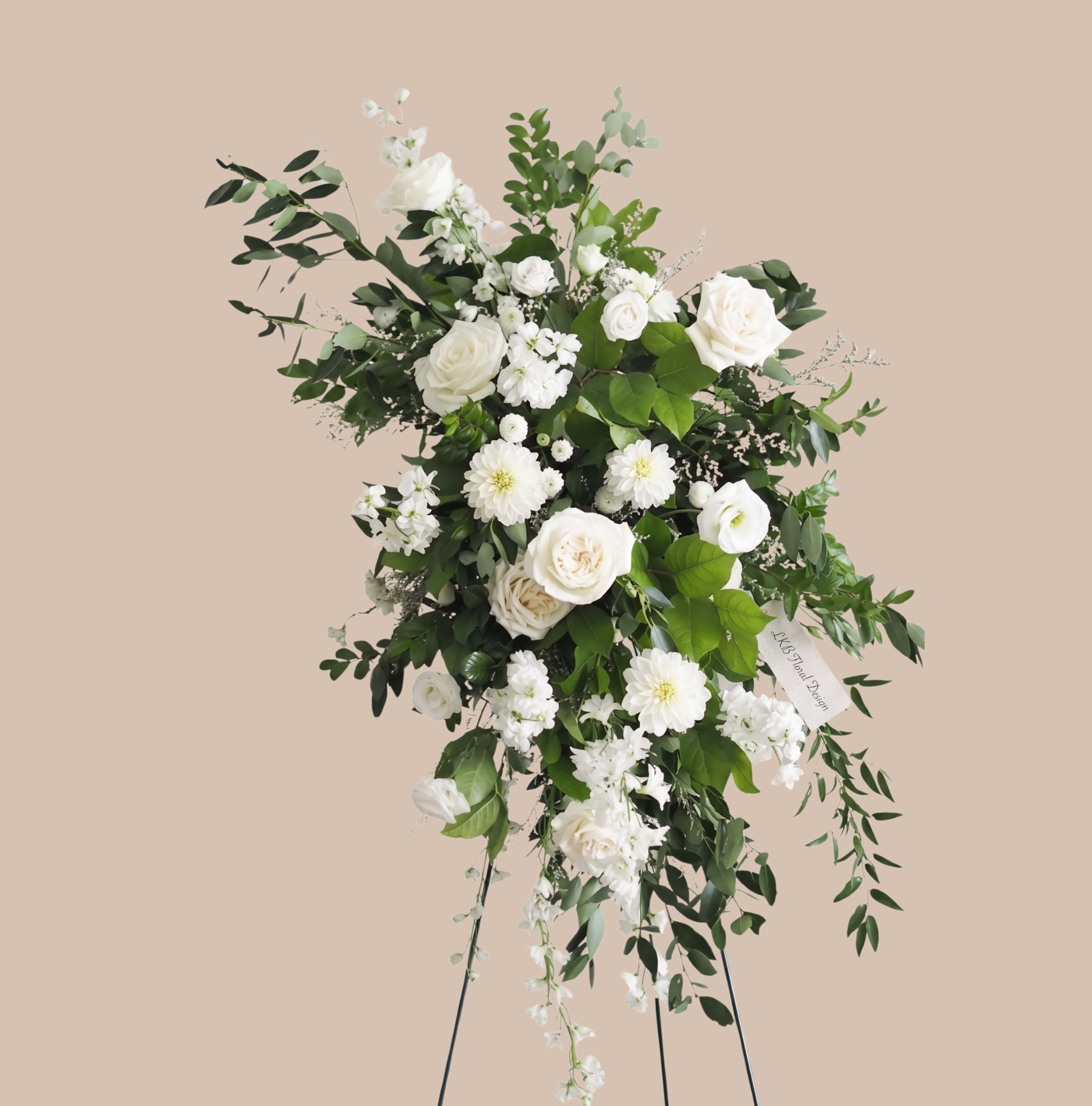 White Meadow Easel - A cascading sweep of white roses, garden roses, lisianthus and daisy-like blooms creates the calm, ceremonial impression of this large standing spray - perfect as a sympathy arrangement or for a memorial service. The White Meadow Spray reads fresh and serene: the soft whites set against abundant mixed greenery give the person you're honoring a quiet, dignified moment of beauty that feels both comforting and respectful.  Arranged on a floral stand in an asymmetrical, flowing silhouette, the spray pairs clusters of pristine roses and softer garden roses with airy lisianthus and cheerful daisy-shaped flowers, finished with trailing greenery for gentle movement. The restrained white-and-green palette and textured foliage make an elegant visual statement; a ribbon tag is included so you can add a brief tribute to accompany the display.  Designed to stand prominently at a service or graveside, this tall arrangement is sized for presentation and immediate impact. It's arranged to travel and arrive ready to display, offering an immediate, comforting presence for family and friends during a difficult moment.