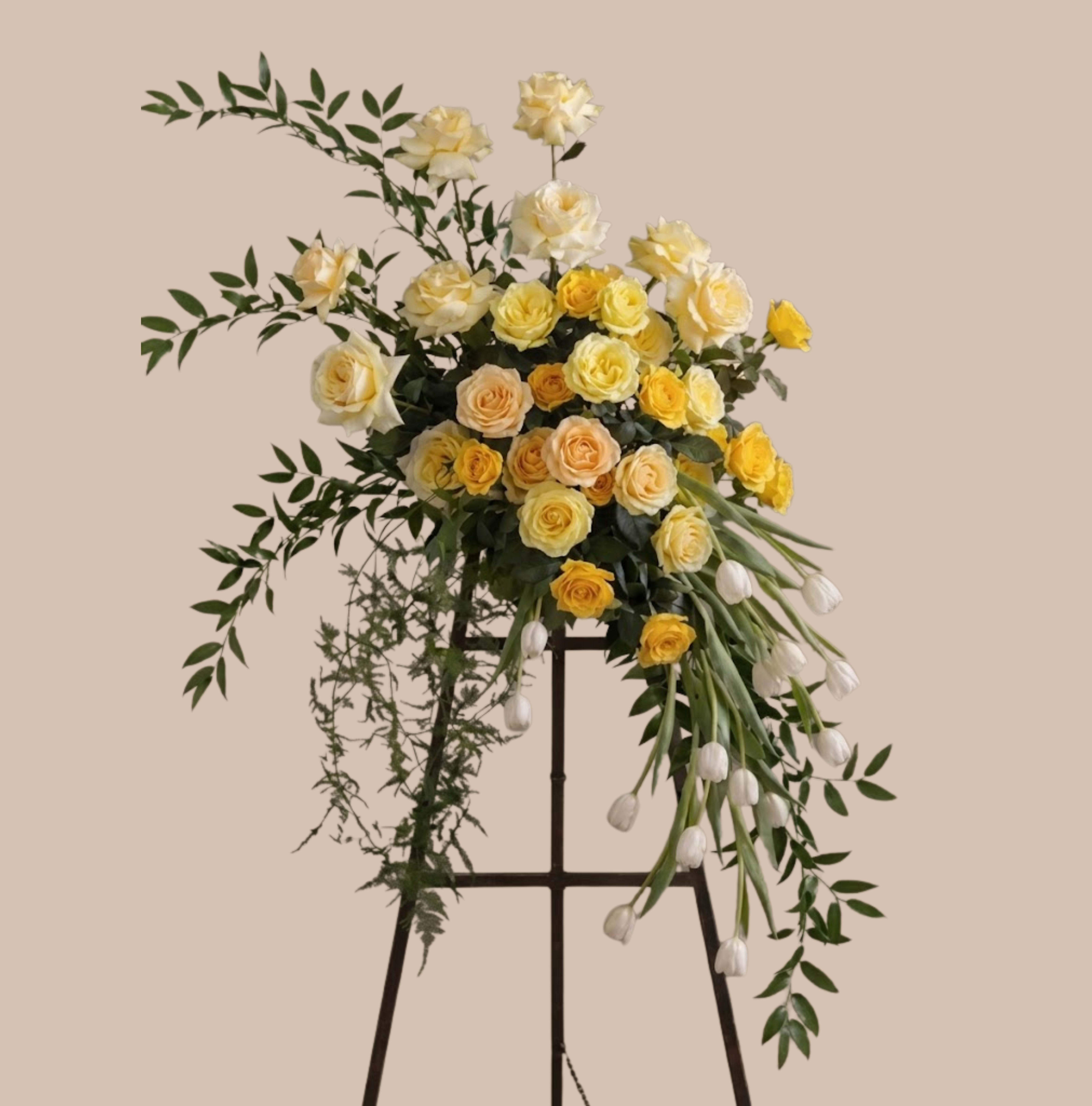 Bonds of Gold Easel - An elevated display of soft pastels and graceful movement, this flower arrangement is a formal easel spray perfect for sympathy tributes or for marking a heartfelt celebration. The vertical presence of the black metal easel and the large asymmetrical spray make it an immediate focal point-an arrangement that reads as elegant and serene, designed to convey comfort or quiet admiration to the person you're honoring.  Cream, yellow, peach roses form the heart of the composition, accented by long, trailing white tulips and airy green foliage that add texture and a gentle sense of motion. The cascading tulips soften the silhouette while the mixed rose tones create warm highlights against the fresh greenery. Mounted on a sturdy black metal easel, the piece offers a polished, professional presentation suitable for a memorial service, a remembrance display, or a refined celebration where a standing arrangement is called for.  Choose this easel spray when you want the recipient to feel truly seen-whether expressing sympathy, respect, or warm congratulations. The vertical scale and durable easel make delivery and display straightforward; please contact us for size options or placement guidance to ensure the arrangement fits the venue and the moment.