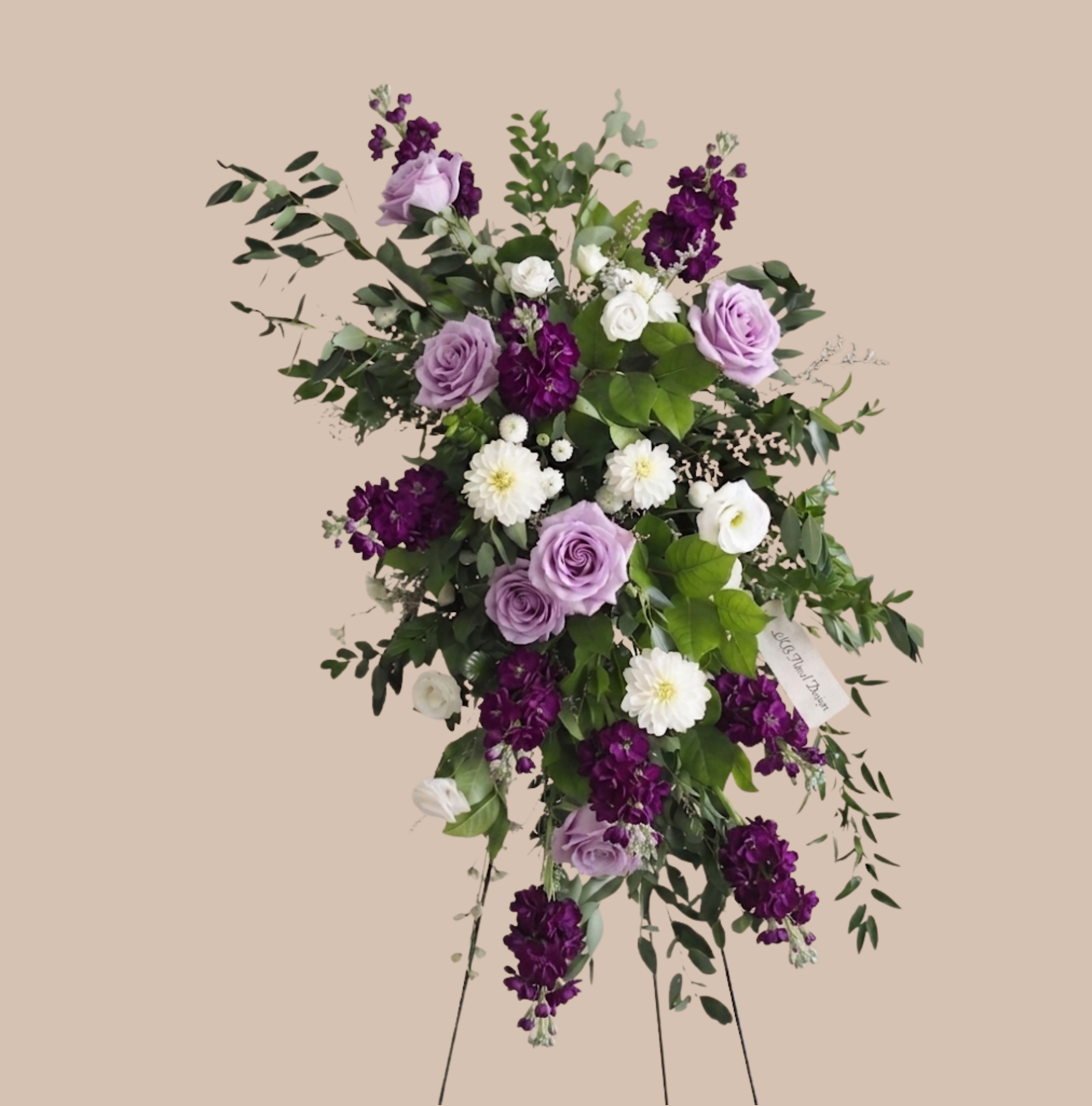 Lavender Symphony Easel - A graceful standing spray that reads like a soft musical phrase, this funeral flower arrangement pairs lavender roses and pale lisianthus with white daisy-like blooms and deep purple accents to create a soothing sympathy tribute. Designed for memorial services and bedside remembrances, the Lavender Symphony Easel offers an elegant, calming presence that honors someone's life with quiet beauty.  The arrangement sits on a sturdy standing easel and features a loose, cascading silhouette with layered greenery for a natural, slightly asymmetrical profile. Garden roses and spray roses mingle with stock and ranunculus textures, producing visual depth-lavender, white, and rich purple hues balanced by fresh green foliage. The composition is airy yet full, large enough to be a focal point at a service while remaining refined and respectful.  Created to convey comfort and remembrance, this spray is intended for family tributes and funeral viewings-the kind of arrangement that helps the recipient feel seen and honored. Sizes and exact flower counts vary by availability; please contact us for personalization requests, delivery timing to services, or to discuss specific color preferences.