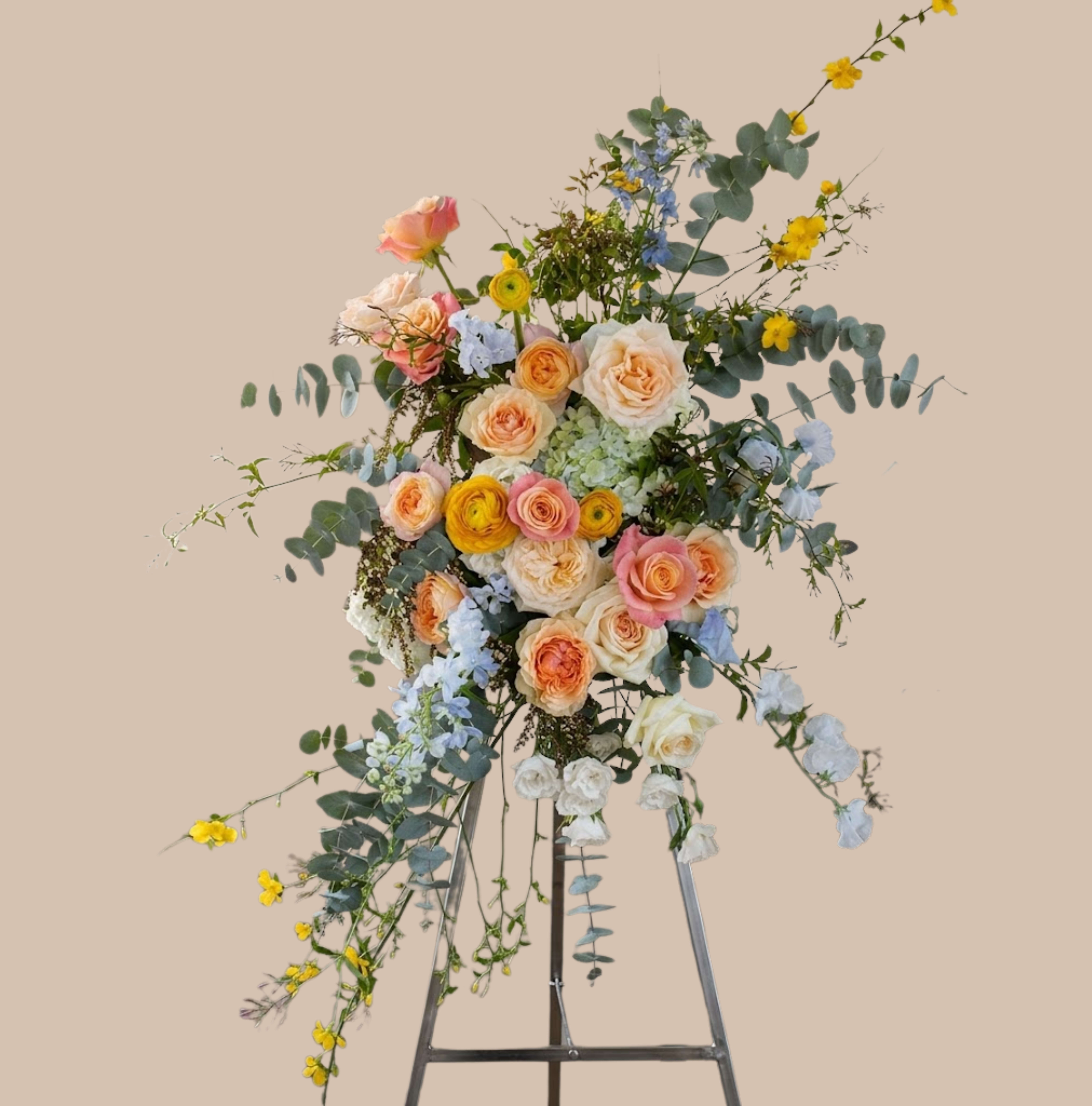 Pastel Clouds Easel - Soft, airy pastels rise from a metal easel stand in this sympathy and funeral flower arrangement that reads like a gentle cloud of comfort. The Pastel Clouds Easel is a tall, standing floral tribute crafted for moments when you want the person you're honoring to feel seen-quietly graceful for a memorial service yet light enough to bring calm to a bedside or entryway.  Clusters of peach, blush, and white roses and garden roses mingle with yellow ranunculus, pale blue delphinium, and light blue hydrangea, while long branching stems and silver-green eucalyptus give the design a loose, cascading silhouette. The mix of lisianthus, spray roses and delicate ranunculus adds layered texture and a soft fragrance; overall the palette of pink, lavender, blue, peach and touches of bright yellow creates a hopeful, springlike impression without feeling seasonal.  Appropriate for sympathy, funerals, or as a thoughtful standing tribute to mark remembrance, this arrangement arrives ready on a sturdy metal easel stand and is scaled to read beautifully in a sanctuary or reception area. Arrange delivery details at checkout; to keep blooms looking their best for services, place away from direct heat and follow simple care instructions provided with the arrangement.
