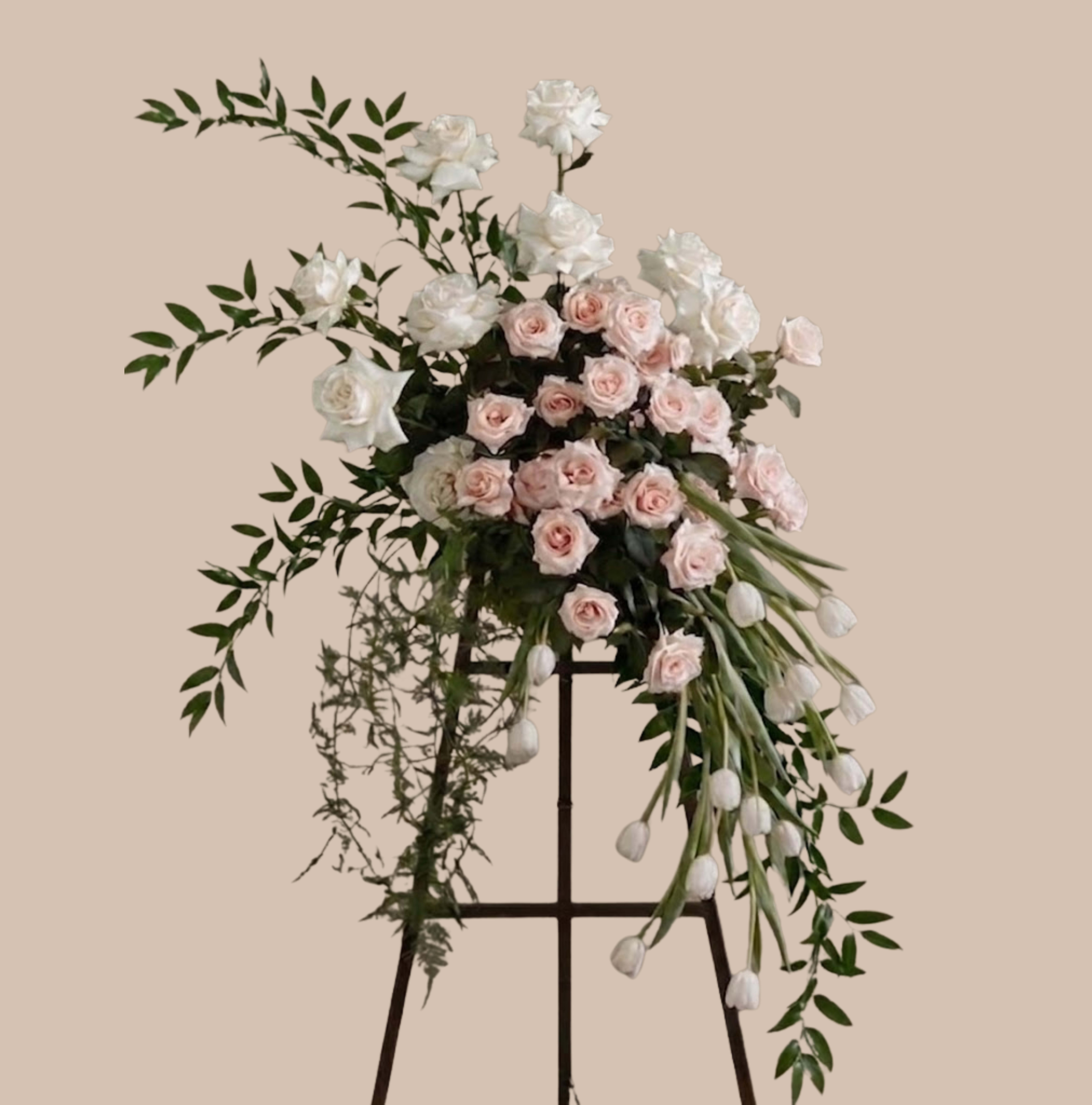 Blush Elegance Easel - A tall, cascading sympathy or celebration arrangement that reads as soft and refined: the blush and white floral cascade on a metal easel stand makes a graceful statement for a wedding, memorial service, or any occasion that calls for elegant vertical impact. The design places pale pink roses and larger white blooms at the visual heart, while trailing white tulips and long, arching greenery create sweeping movement that draws the eye from top to bottom - the kind of piece that helps the person you're honoring feel beautifully remembered.  This easel display mixes clusters of delicate pale pink roses with prominent white blossoms and elongated white tulips, set against loose, airy green foliage for a natural, garden-fresh texture. The metal easel stand gives the arrangement formality and height, making it suitable for aisles, ceremony backdrops, reception entrances, or a distinguished everyday display. Designed to be seen from a distance, it balances romantic color with refined structure.  Ideal for commemorating a meaningful moment or celebrating a life event, this arrangement conveys sympathy, love, or congratulations with quiet elegance. For best longevity, keep the flowers in a cool environment away from direct sun and drafts until delivery; sizes and exact bloom counts may vary slightly as each piece is handcrafted to preserve the intended cascading silhouette.
