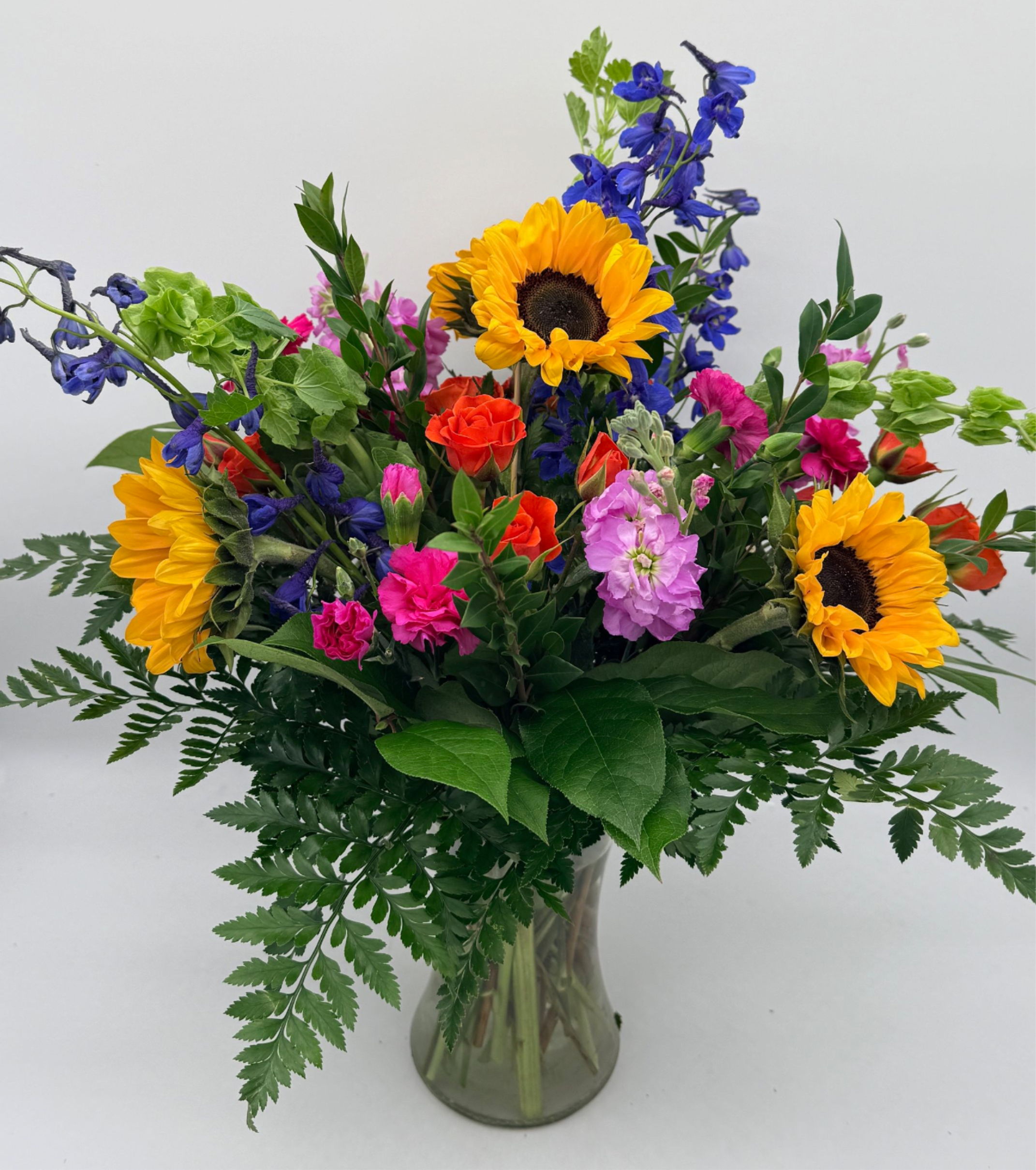 Vibrant mixed bouquet with sunflowers, pink and orange blooms, and blue delphinium in a clear glass vase.