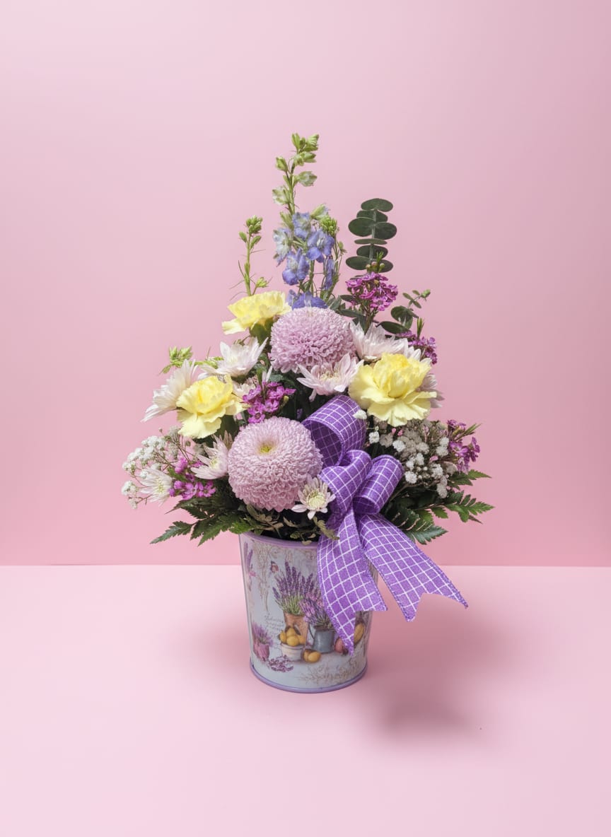Pastel Serenity - Soft, cheerful, and full of gentle charm, Pastel Serenity is a lavender-themed floral arrangement that uplifts and delights. Nestled in a coordinating lavender container, it features fragrant lavender cremones, sunny yellow carnations, delicate pink cushions, and elegant purple larkspur. Accents of pink waxflower and airy baby’s breath add texture and a light, whimsical touch, all finished with a purple-and-white plaid ribbon for a sweet, polished look. Perfect for birthdays, thinking-of-you gestures, or simply bringing a serene pop of color to any space.   All flowers, floral colors, and containers are subject to change due to availability.