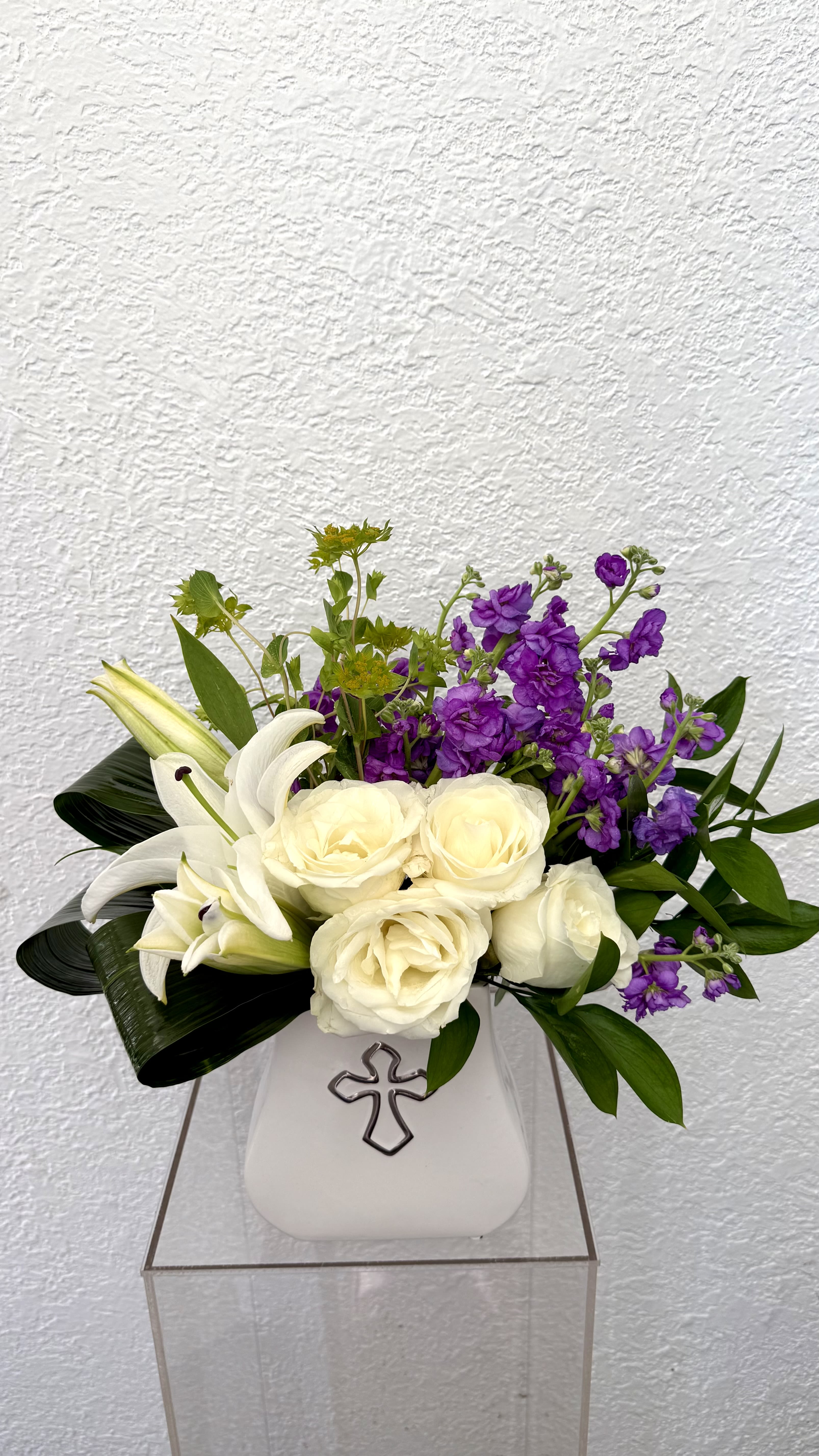 Grace Upon Grace - Designed with graceful white lilies, creamy ivory roses,  and rich purple blooms, this stunning arrangement reflects beauty, faith, and remembrance. Presented in a keepsake cross vase, Grace Upon Grace is a heartfelt gift for sympathy, memorials , encouragement, or spiritual celebrations. 