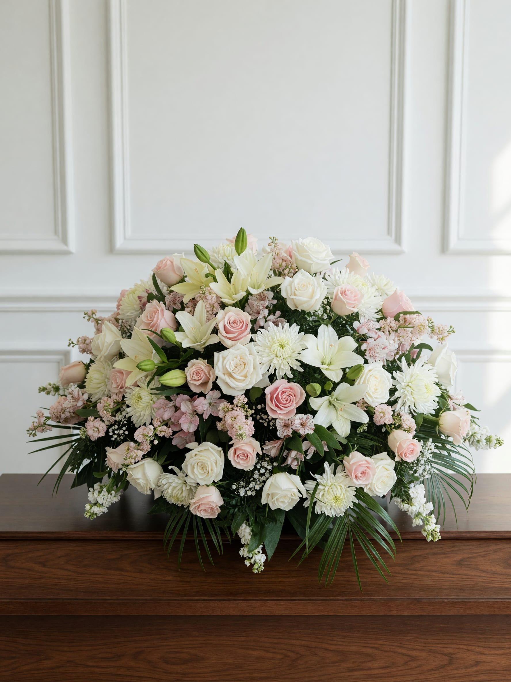 Centerpiece of Hope - A softly layered casket spray in white, blush pink and pale green tones designed to bring a quiet, reverent presence to a funeral or memorial service. This casket spray features roses, lilies, chrysanthemums, hydrangea and daisy-like blooms arranged in a large, low, rounded composition so the person you're honoring will look serenely surrounded by light and gentle color. The pale palette and dense, rounded shape create a refined mood that pairs especially well with "cross of Hope" remembrance for a formal sympathy setting.  Flowers are tightly clustered across a broad base with dark green foliage and long palm fronds extending for contrast and structure, giving the piece an abundant yet composed silhouette suitable for display on a table or casket. The design coordinates beautifully with the matching "Cross of Hope" on a metal easel stand when you want a unified memorial presentation; ribbon accents add a subtle finishing touch. For placement, this large spray is intended for casket or display-table use and is built to read full and elegant from the front of the service.