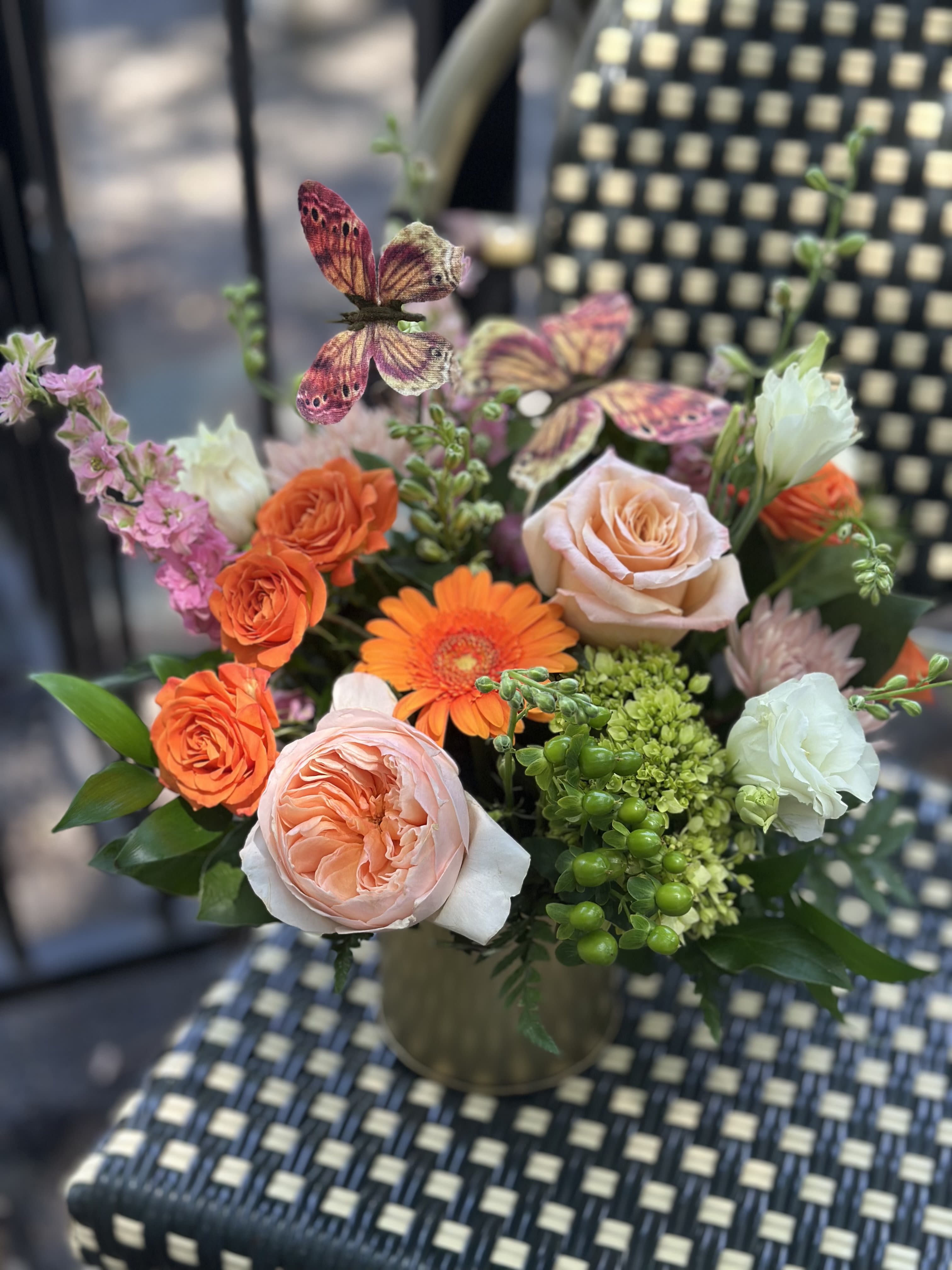 Garden of Wings - A cheerful celebration of color and texture. Garden of Wings is a plucky arrangement that features soft blush roses, vibrant orange blooms, pink accents, and whimsical green accents. Butterfly details flutter above the flowers, adding a touch of fun and make this design feel like a summer garden captured in a vase.