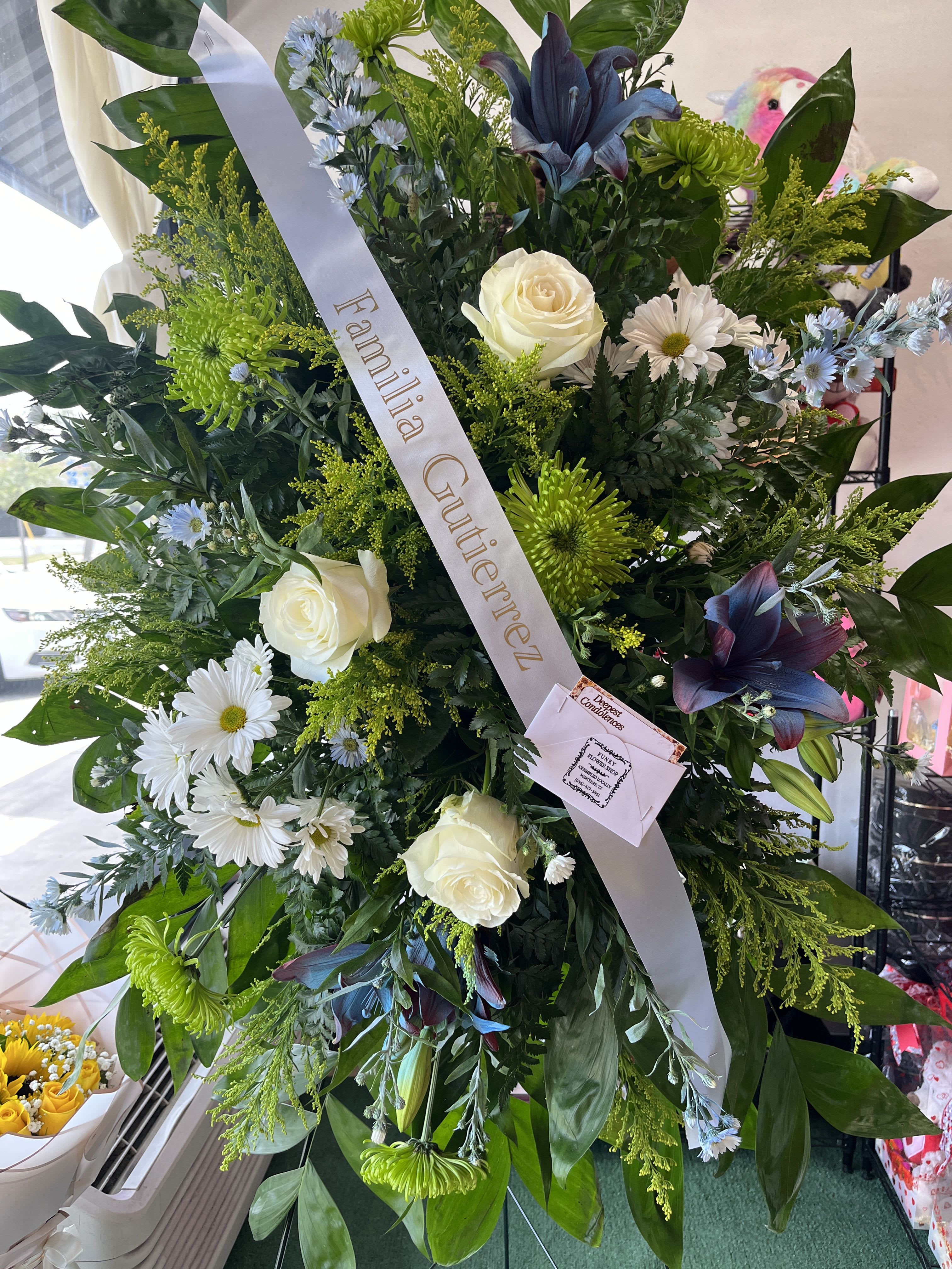 tranquil farewell - A tall, formal standing sympathy arrangement that reads as a respectful and elegant tribute - ideal as a funeral spray or condolence offering. The upright, layered composition centers on white roses and white daisies set against bright green chrysanthemums and deep blue-purple lilies, so the person you're honoring will be remembered with a calm, dignified presence.  This large spray is densely arranged with abundant dark green foliage that gives the design a full, structured silhouette. Finished with a diagonal white or pale satin ribbon (some examples show gold lettering or a personalized print like "Familia Gutierrez"), the piece includes a sympathy ribbon and attached card and is mounted on an easel for display. The white, green, blue, purple, and subtle yellow tones combine to convey serenity and quiet comfort.  For a grieving family member or close friend, this arrangement delivers a solemn, consoling statement appropriate for memorial services and graveside ceremonies. If you'd like the ribbon personalized or need delivery to a service location, please select ribbon text and delivery options at checkout.