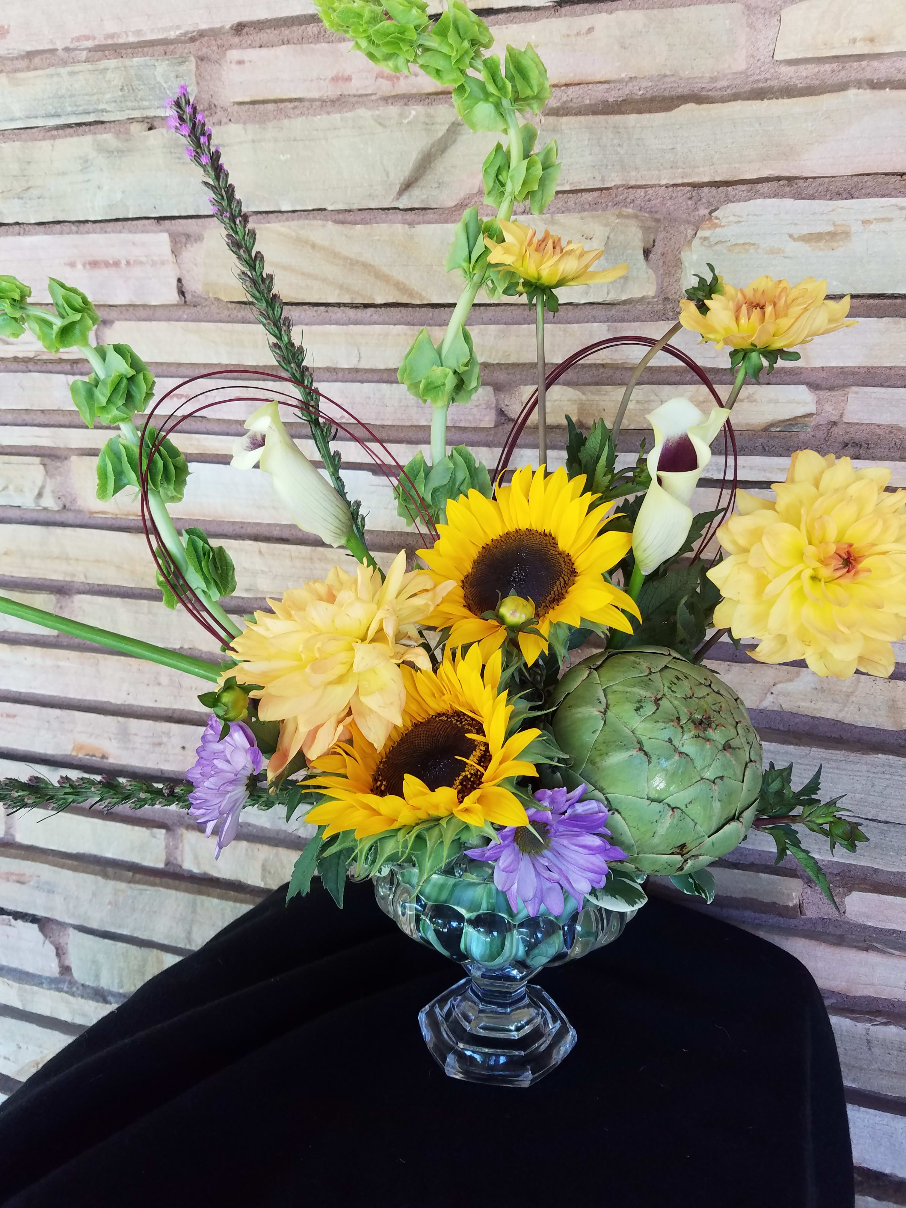 Brooklyn Decker Bouquet - This arrangement contains bells of Ireland, mini callas, sunflowers, liatris, dahlias, accented with an artichoke in a crystal pedestal ( may be substituted )