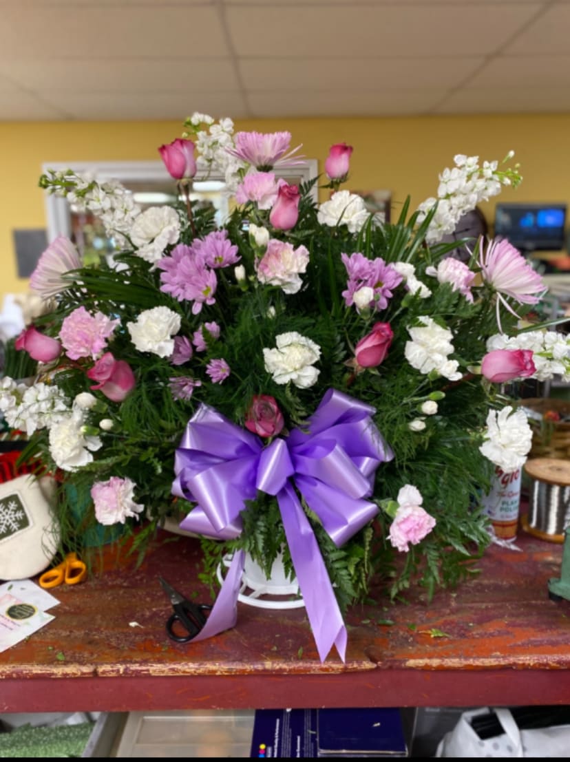 Purple Glory Funeral Basket - Lush shades of pink and white bloom from a generous white basket, tied with a striking lavender-purple satin bow-an ideal birthday bouquet, thank-you gift, or sympathy arrangement that reads both celebratory and comfortingly gentle. The mixed bouquet of roses, carnations, chrysanthemums, and daisies creates a layered, textured silhouette so the person you're thinking of will feel truly seen by its fullness and classic charm.  This large, full design features pink rose buds nestled among white and pink carnations, with soft lavender daisy-like and chrysanthemum blooms rising for added height and interest. Abundant green foliage fills the basket for a traditional florist look, while the purple ribbon provides a bright contrast that highlights the pastel palette. The rounded, slightly vertical shape makes it a strong presence on a table or memorial stand.  Perfect for sending warm congratulations, expressing heartfelt thanks, or offering comfort, this arrangement arrives ready to display in its white basket-no additional vase needed. Care: keep blooms in a cool spot away from direct sun and refill the basket's water as needed to maintain freshness.