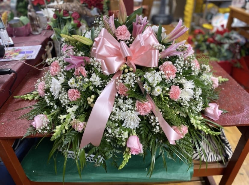 Pink Pasture with Baby Breath with Pink Bow Casket Spray  - A soft, full casket spray in pink, white, and green creates a gentle, comforting display for a funeral or memorial service. This funeral arrangement balances the formality of a casket spray with a tender, celebratory feel-appropriate for honoring a life at a funeral, memorial, or celebration of life while also suitable for marking a milestone like Mother's Day or an anniversary remembrance.  Dense clusters of pink lilies, pale carnations, and small daisy-like blooms are artfully layered into a low, rounded shape that reads lush from every angle. Sprays of greenery soften the edges and add contrast, and a prominent pink satin ribbon tied at the center gives the piece a refined, gift-like finish. The palette of blush pinks and creamy whites lends a quietly romantic, thoughtful tone without being flashy.  Designed to lie gracefully atop a casket, this arrangement is sized to provide full coverage and visual impact while remaining elegant and understated. It's the kind of tribute that helps the person you're remembering feel honored and seen; allow our team to handle delivery and placement so your focus can stay on celebrating their life.