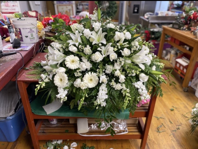 Glorious Morning Casket Spray - White lilies and white roses from the heart of this Glorious Morning casket spray, a large sympathy flower arrangement designed to honor and comfort. As a casket spray for a funeral or memorial service, it reads serene and formal-the kind of design that lets the person you're remembering feel beautifully respected and seen.  This arrangement is built with layered white lilies, white daisy-like blooms, pale pink roses, carnations and stock, arranged densely over floral foam to create a full, rounded silhouette. Lush greenery fills and cascades from the edges, giving texture and a gentle flow along the sides of the spray. The substantial size and close-set blossoms make it suitable as a centerpiece for a service or to rest atop a casket.  For someone planning a meaningful farewell, this spray expresses sympathy with quiet elegance. It arrives ready to place on the service table or casket, assembled on a work table and secured in floral foam for lasting freshness-perfect when you want a respectful, long-lasting tribute.