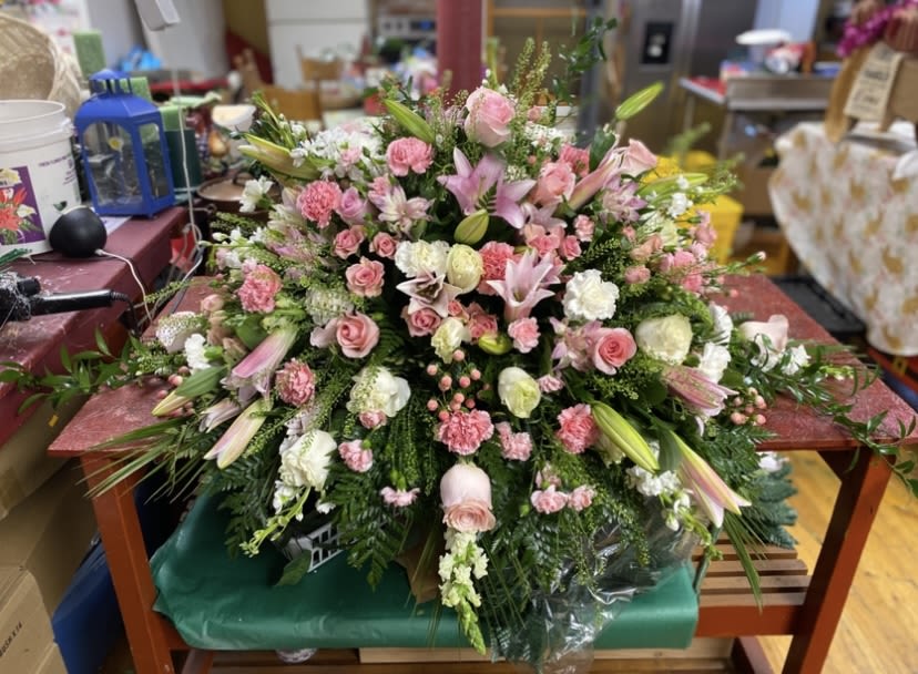 Strawberry And Cream Casket Spray - A lush, low casket spray that reads as soft and celebratory-perfect for honoring a loved one with gentle pinks and creams. This flower arrangement for funeral or memorial services pairs lilies, roses, carnations, alstroemeria, and daisies in a sprawling, formal design meant to rest gracefully atop a casket or serve as a centerpiece at a remembrance gathering. The palette of pink, white, green, and touches of purple creates a comforting, romantic tribute that helps the person you're remembering feel honored and truly seen.  The composition is densely layered over rich greenery for a full, abundant look: large lilies and open roses punctuate the plane, carnations add texture and body, while alstroemeria and daisies provide delicate, eye-catching accents. Finished with discreet plastic wrap for transport and placement, the arrangement is broad and low-designed to present a continuous cascade of blooms without obstructing view during services. Its scale and formal styling make it an appropriate choice for a meaningful funeral or memorial display.
