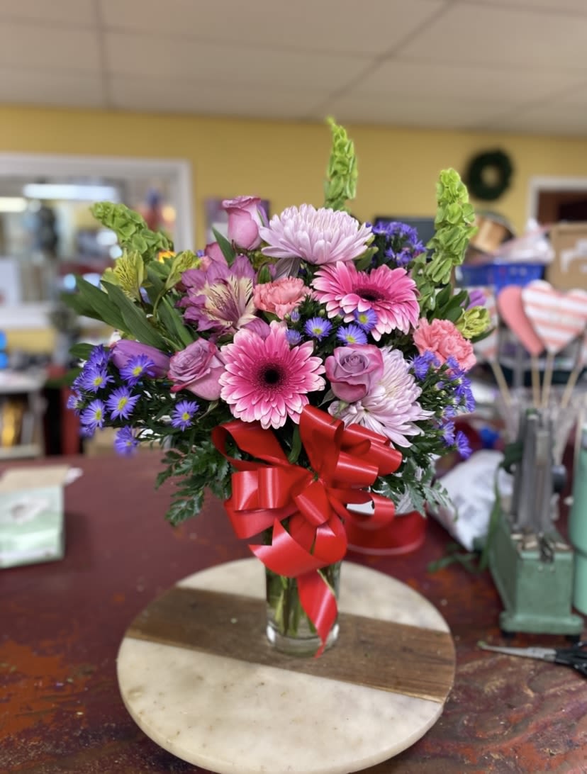 Shining Beauty - A cheerful burst of pinks and purples, Shining Beauty is a birthday bouquet and romantic flower arrangement that makes the recipient feel truly celebrated. This hand-tied mix centers on bold gerbera daisies and classic roses, with carnations and alstroemeria adding soft, ruffled texture-perfect for saying happy birthday, thank you, or I'm thinking of you in a bright, uplifting way.  The clear glass vase shows off dense, layered blooms in shades of pink, purple, red, and white, tied with a large red ribbon for a festive finish. Chrysanthemums and aster-like daisy fillers bring light, airy contrast while bright green accent spikes add height and lively contrast to the softer petals. The design reads full and abundant, offering visual depth and a variety of textures that catch the eye from every angle.  Ideal for celebrating a friend, partner, or family member, this arrangement sends a message of warmth and joy the moment it's set on a table. Vase-ready and hand-tied for an effortless display, keep flowers fresh by changing the water regularly and trimming stems slightly every few days to prolong the bloom life.