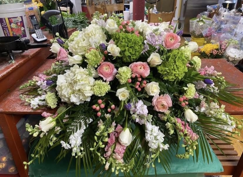 Enchanted Garden Casket Spray - Soft pastel blooms arranged in a broad, cascading shape create a peaceful, reverent presence for a funeral or sympathy setting. This casket spray is designed as a traditional, low, expansive arrangement meant to rest across a casket - a comforting way to honor the life of the person you're remembering and to let loved ones feel supported in a tender moment.  White hydrangeas form the lush foundation while pale stock and white tulips add vertical softness; pink ranunculus bring delicate layers of petal texture and gentle color. Clusters of green blossoms and dark foliage provide contrast that makes the pastels read with quiet depth. The overall effect is formal and abundant, with a cascading silhouette that drapes naturally across the casket for full coverage and graceful lines.  For families seeking a respectful tribute, this spray conveys sympathy and heartfelt remembrance without overpowering the service. Its low profile and broad spread are sized to sit directly on the casket; please contact us with the casket dimensions or special requests so we can ensure a perfect fit and freshest seasonal blooms.