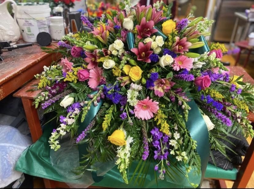 Mystical Garden Casket Spray - A lush cascade of pink lilies, purple accents, and sunlit yellow and white roses-this large mixed bouquet makes a joyful statement for a birthday bouquet or celebratory gift. The rounded, cascading shape reads as abundant and energetic, the kind of arrangement that will make the person you're thinking of feel truly celebrated and seen.  This dense bouquet layers pink gerbera daisies, fragrant lilies, classic roses, smaller spray roses, alstroemeria and stock for texture and movement, with additional purple filler blooms to deepen the palette. Teal ribbon and green floral wrap provide a striking contrast to the soft florals and greenery, giving the arrangement a polished, upscale look. It's substantial in size and volume, meant to fill a room with color and scent.  Perfect for birthdays, anniversaries, or to mark a big congratulations, this arrangement suits someone who loves bold color and lush presentation. To keep the blooms fresh, place in clean water and recut stems if possible; if you need delivery or special timing, choose your preferred delivery date at checkout so the bouquet arrives at its peak.