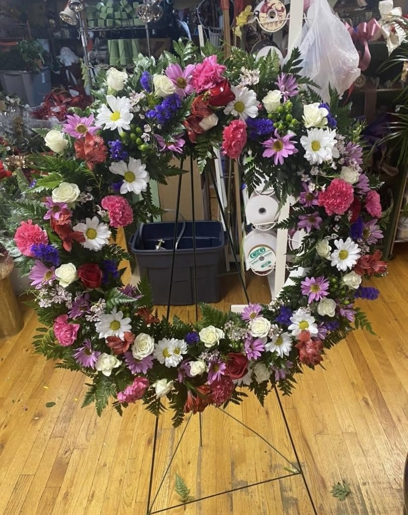 Endless Love Fresh Wreath on Easel - A heart-shaped sympathy wreath that speaks of enduring affection, the Endless Love Fresh Wreath on Easel is a large floral tribute ideal for memorials, funerals, or a heartfelt display at a celebration of life. The heart form and rich color mix-soft pinks, pure whites, romantic reds and touches of purple-send a clear message of love and remembrance the person you're honoring will be remembered and cherished.  This arrangement combines roses, daisies, carnations, alstroemeria and other seasonal blooms set against dense, fern-like greenery that outlines and layers the heart for a full, textured look. Mounted on a sturdy metal easel stand, the wreath reads clearly from a distance and creates an elegant focal point; the mixed blooms provide variety in shape and scent while the color palette balances tenderness with dignity.  Perfect for sympathy services, memorial displays, or as a loving tribute at a wedding remembrance, this wreath conveys compassion without words. The large scale and professional easel presentation make placement simple-order with confidence for delivery timed to the service so the person you're honoring is remembered exactly as they deserve.