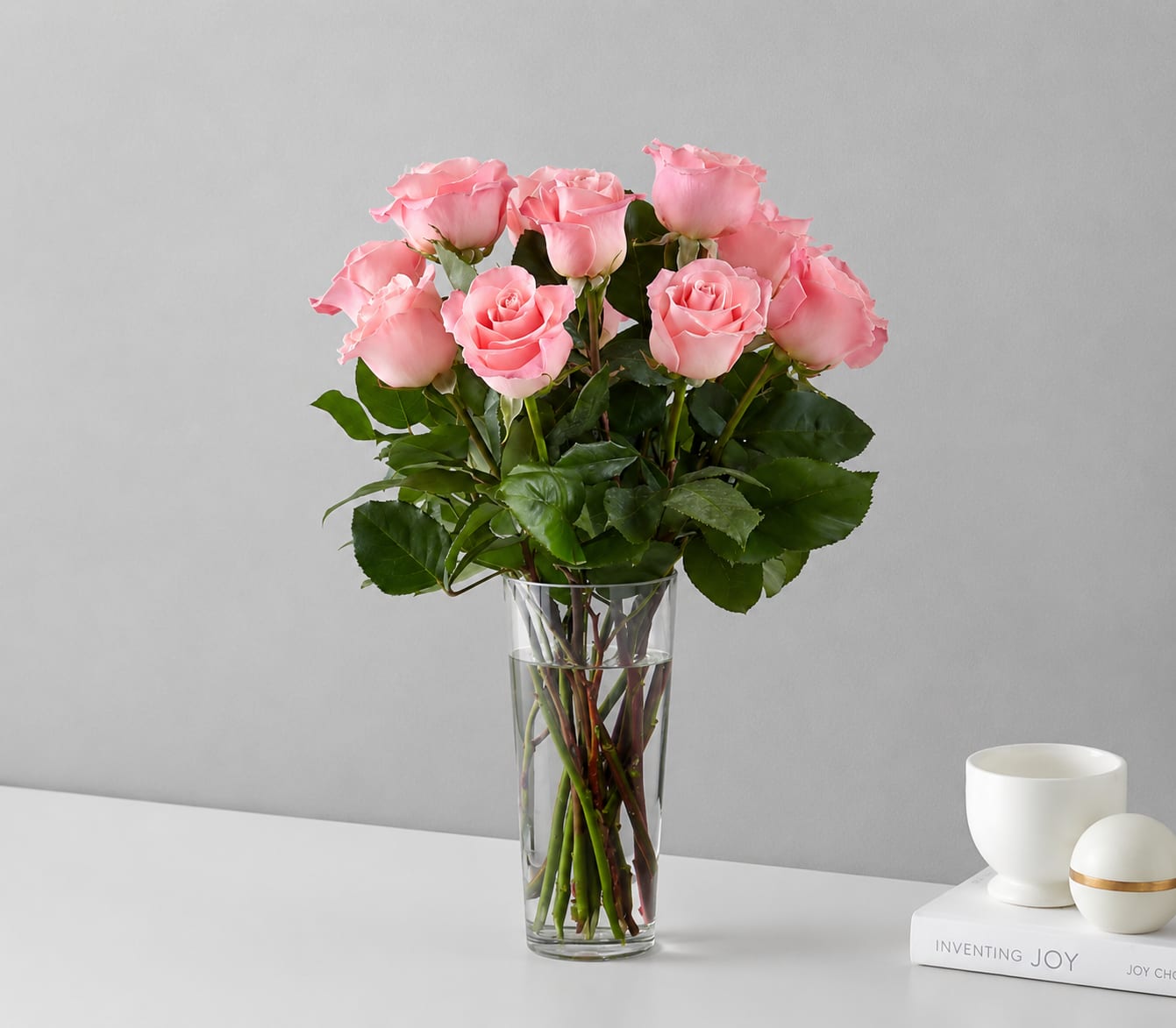 Pink Roses - Soft pink roses gathered tall in a clear glass vase create a romantic, garden-fresh statement for anniversaries, birthdays, or the everyday moment when you want someone to feel truly cherished. This pink roses flower arrangement reads feminine and celebratory-perfect for love and romance, a new baby welcome, a thoughtful just-because surprise, or a comforting sympathy gift-while also fitting seasonal moments like Valentine's Day or Breast Cancer Awareness Month.  Full, rounded pink rose blooms are paired with filler flowers of choice and abundant dark green foliage for a lush, airy silhouette. Long stems are visible through the vase for a clean, elegant look. The design arrives ready to display and conveys warmth and care the moment it's set on a bedside table, mantle, or hospital room.  This bouquet is meant to make the recipient feel seen and celebrated-whether you're saying happy birthday, get well soon, I love you, or offering gentle sympathy. For best enjoyment, trim stems and refresh the water every few days to extend the life of the roses.