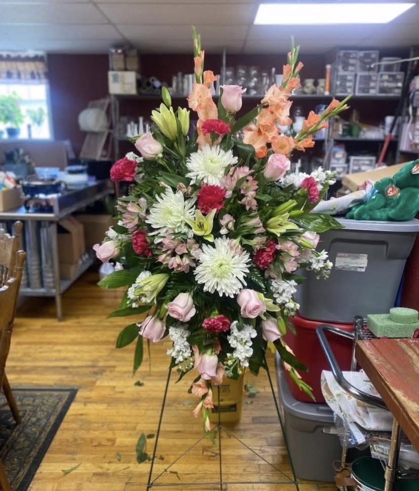 Splendid Sunrise Standing Spray - Soft pink roses and airy white chrysanthemums form the heart of this tall standing spray, making an immediate, uplifting impression for a memorial or sympathy arrangement. The Splendid Sunrise Standing Spray reads as a respectful tribute-a formal, upright floral statement that still feels warm and hopeful, perfect for honoring someone special or marking a ceremonial occasion.  Peach gladiolus and clusters of lilies, alstroemeria, and carnations add vertical drama and soft fragrance, while deep green foliage grounds the dense, layered composition. Long stems rise above the main mass and a gentle cascading lower section gives the arrangement balanced movement; finished on a metal easel with a yellow container, it presents with dignified height and presence.  Choose this spray when you want the person you're thinking of to feel remembered and celebrated with tenderness-the colors convey comfort without somberness. The standing design arrives ready to display on its included easel; for best longevity, keep the location cool and out of direct sun and follow any included care instructions to preserve the blooms during memorial services or viewings.