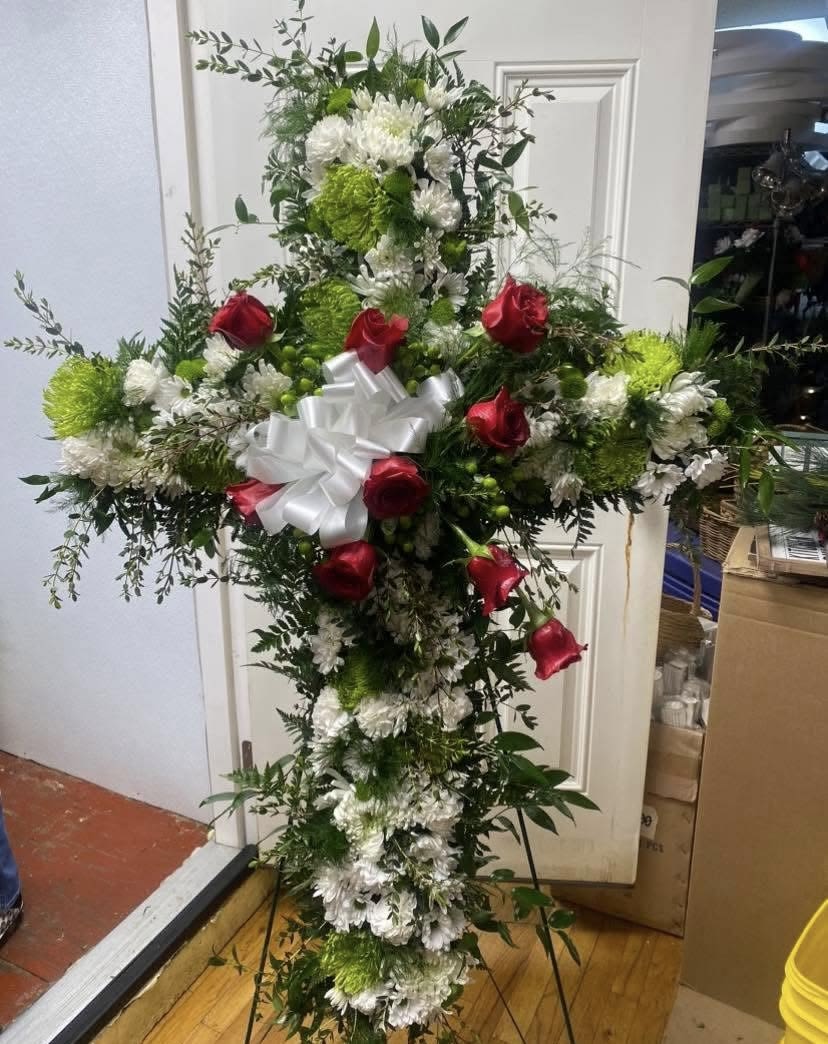 Grace Everlasting Fresh Cross on Easel - A dignified standing cross crafted from dense white blooms and bright green pompon chrysanthemums, accented with a few red roses-this sympathy flower arrangement brings a solemn, devotional presence to a memorial or funeral service. The white ribbon bow at center and the upright easel present the cross with formal grace, making it an appropriate tribute when you want the person you're honoring to feel remembered and respected.  White roses and chrysanthemums form the body of the cross, with green pompons adding subtle color contrast and texture while red roses are placed across the front for a touch of warmth and meaning. Airy greenery fans gently around the edges to soften the silhouette and create a balanced, symmetrical composition. The cross is mounted on a standing easel and finished with a large white ribbon for a traditional, ceremonial look.  Perfect for sympathy, funeral services, or memorial dedications, this arrangement communicates reverence and lasting remembrance. Available in a standard standing size suitable for chapel displays; place in a cool location out of direct sun until the service and keep floral foam moist for best freshness.
