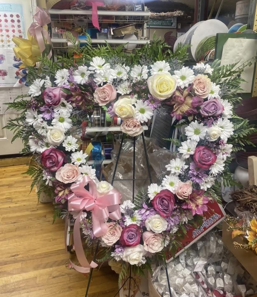 Gracious Blooms Open Heart on Easel - A large heart-shaped wreath of roses and daisies creates an immediate, sentimental impression-perfect for sympathy, remembrance, or expressing deep love. This heart-shaped sympathy arrangement is designed to console and honor: the soft palette of pink, lavender, cream and white roses paired with white daisies reads gentle and reverent, so the person you're remembering will feel truly seen and comforted.  The wreath is full and layered, built with mixed roses, white daisy blooms, alstroemeria accents and small purple filler flowers, all cushioned by airy greenery that defines the heart silhouette. A pink satin bow at the lower left adds a tender finishing touch, and the piece is presented on a sturdy black metal easel for prominent display at a service or memorial.  Choose this heart wreath when you want to send a message of love and remembrance-a respectful, heartfelt tribute that reads both romantic and consoling. Because it arrives on an easel and is sized for display, it's ready for placement at a ceremony; no extra assembly is required.
